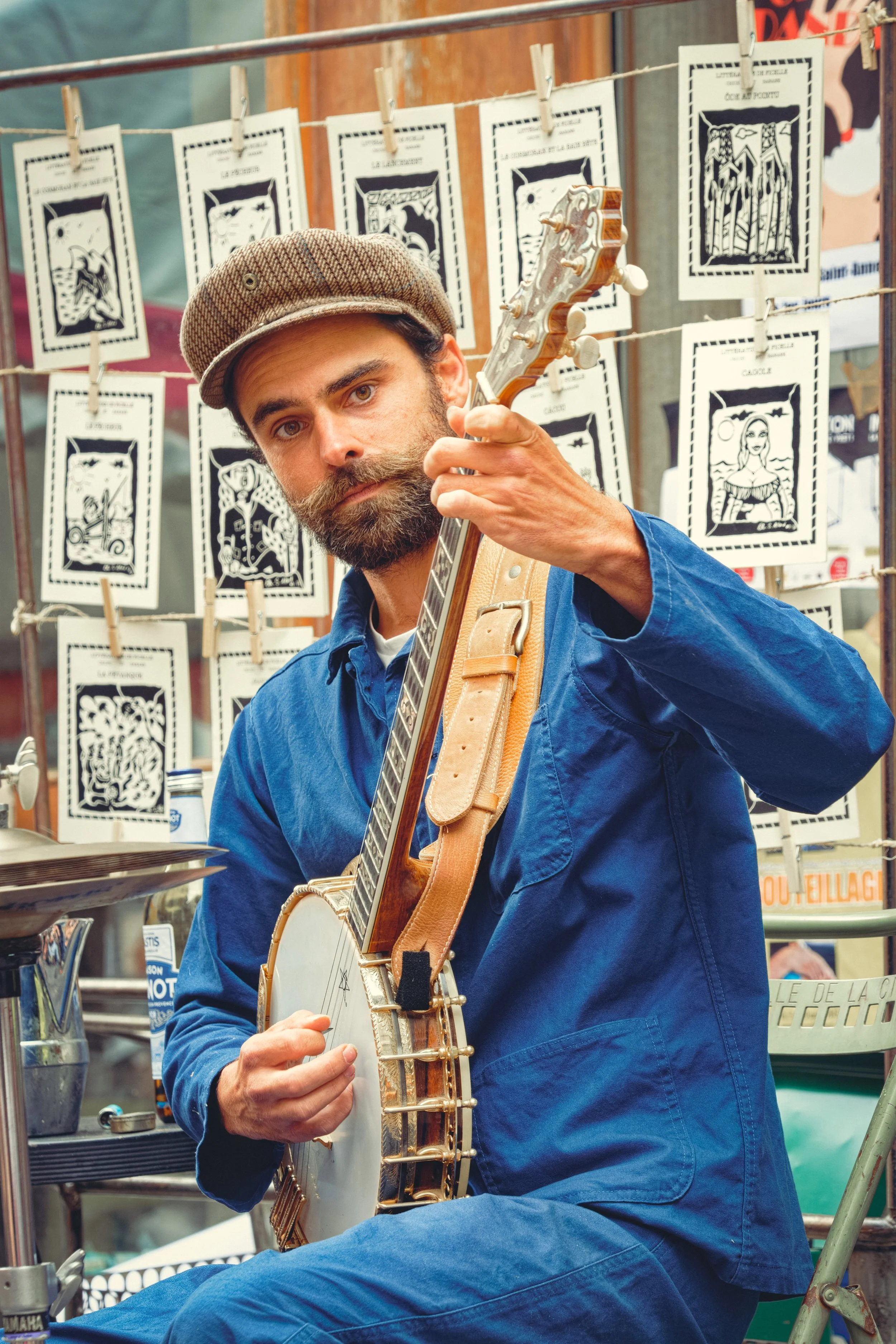 Homme en salopette bleue et casquette plate jouant du banjo, avec des impressions en noir et blanc exposées sur une ligne en arrière-plan.
