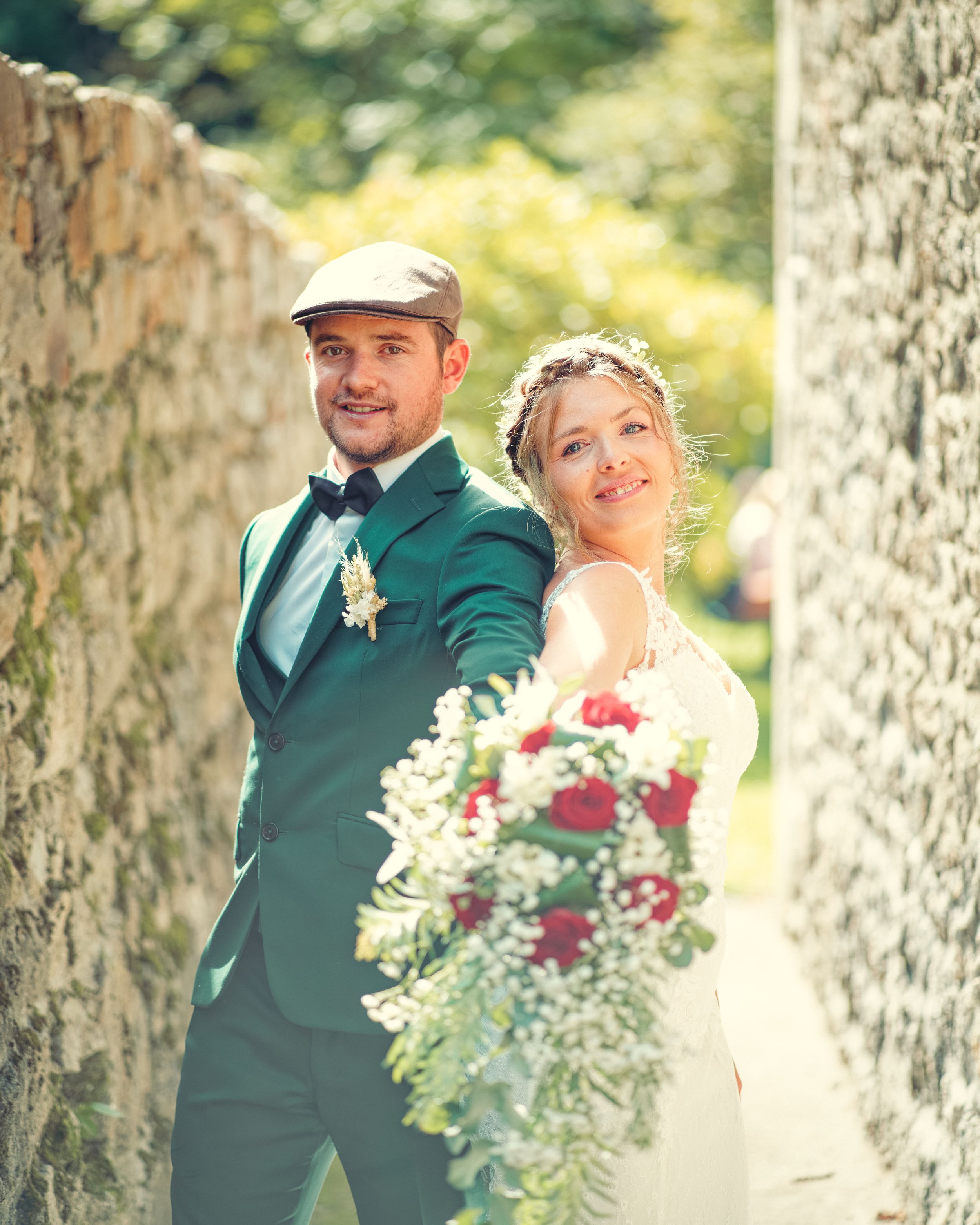 Mariée en robe blanche tenant un bouquet à côté du marié en costume vert et casquette, se tenant entre des murs de pierre.