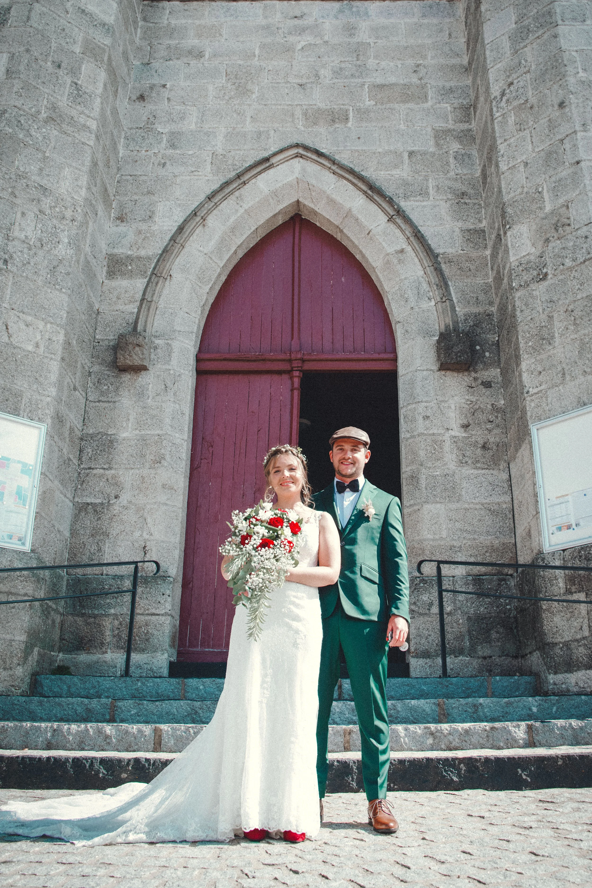 Bride and groom standing outside a stone building with a wooden door, holding flowers.