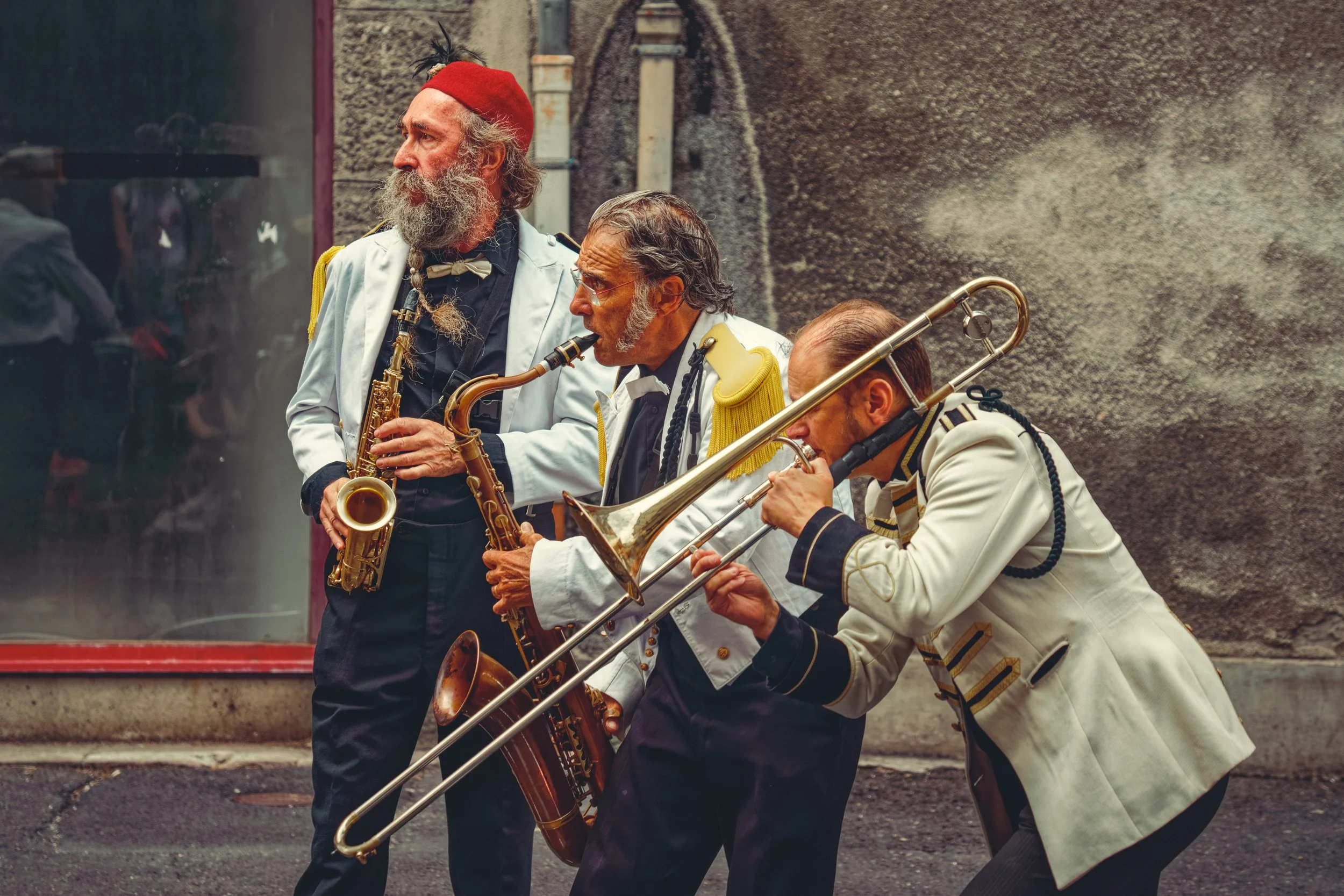 Un trio de musiciens de rue jouant d'instruments en cuivre. L'un porte un fez et joue du saxophone, un autre, en uniforme de cérémonie, joue d'un deuxième saxophone, et le troisième musicien joue du trombone. Ils sont à l'extérieur, près d'un bâtiment aux murs à la texture rugueuse.