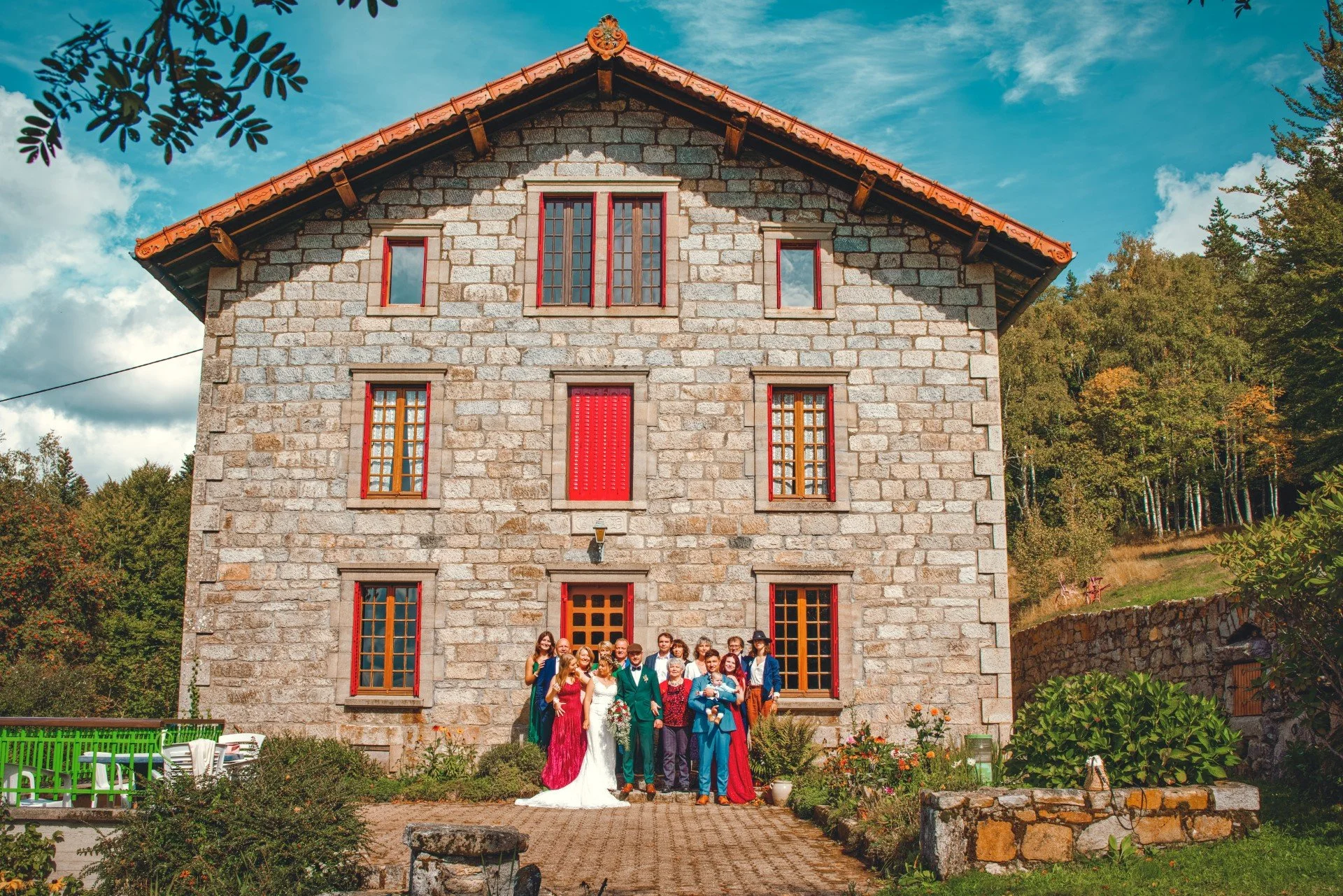 Un groupe de personnes posant devant une grande maison en pierre avec des volets rouges, entourée de verdure et de fleurs, sous un ciel bleu.