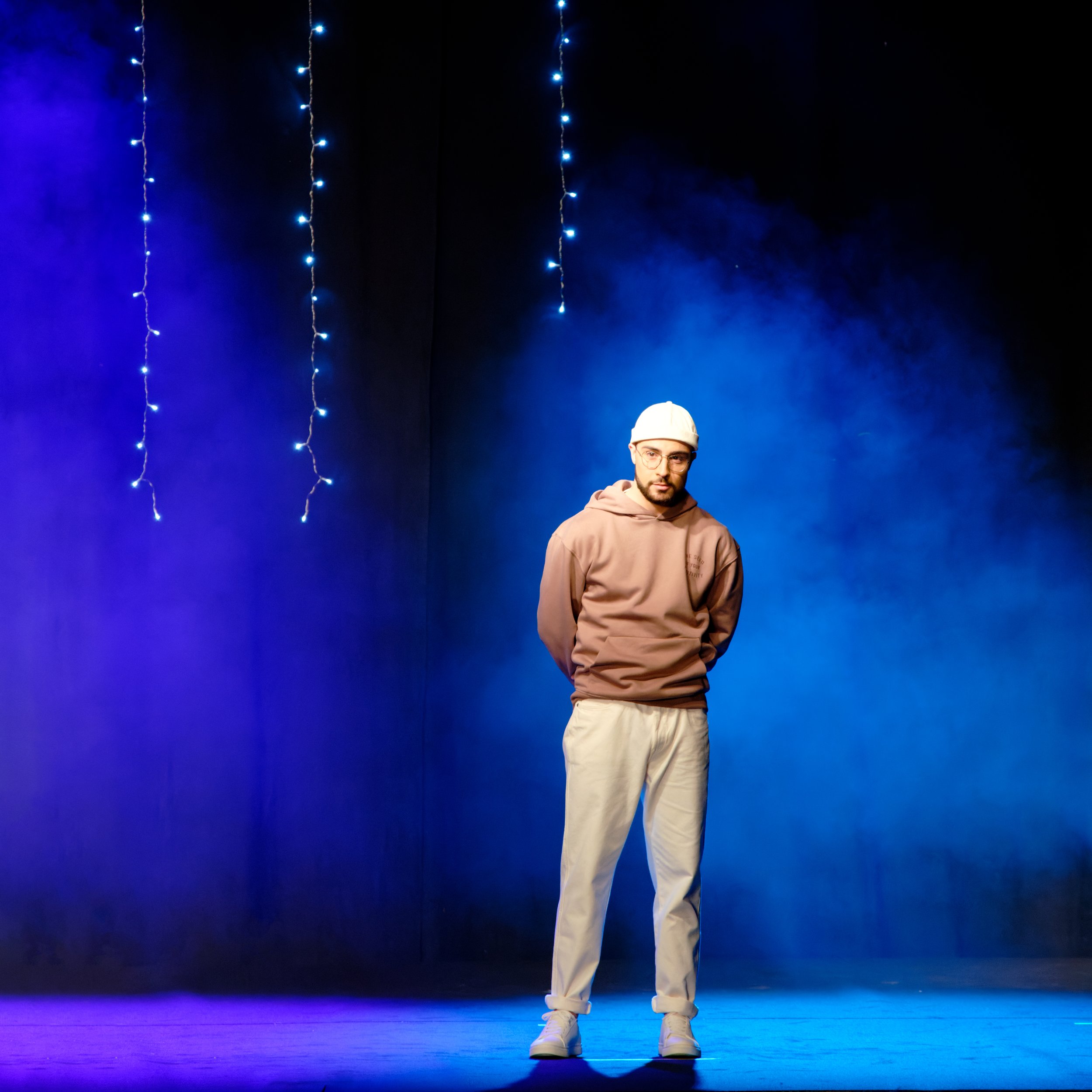 Personne en sweat à capuche debout sur une scène avec un éclairage bleu et violet, entourée de guirlandes lumineuses suspendues.