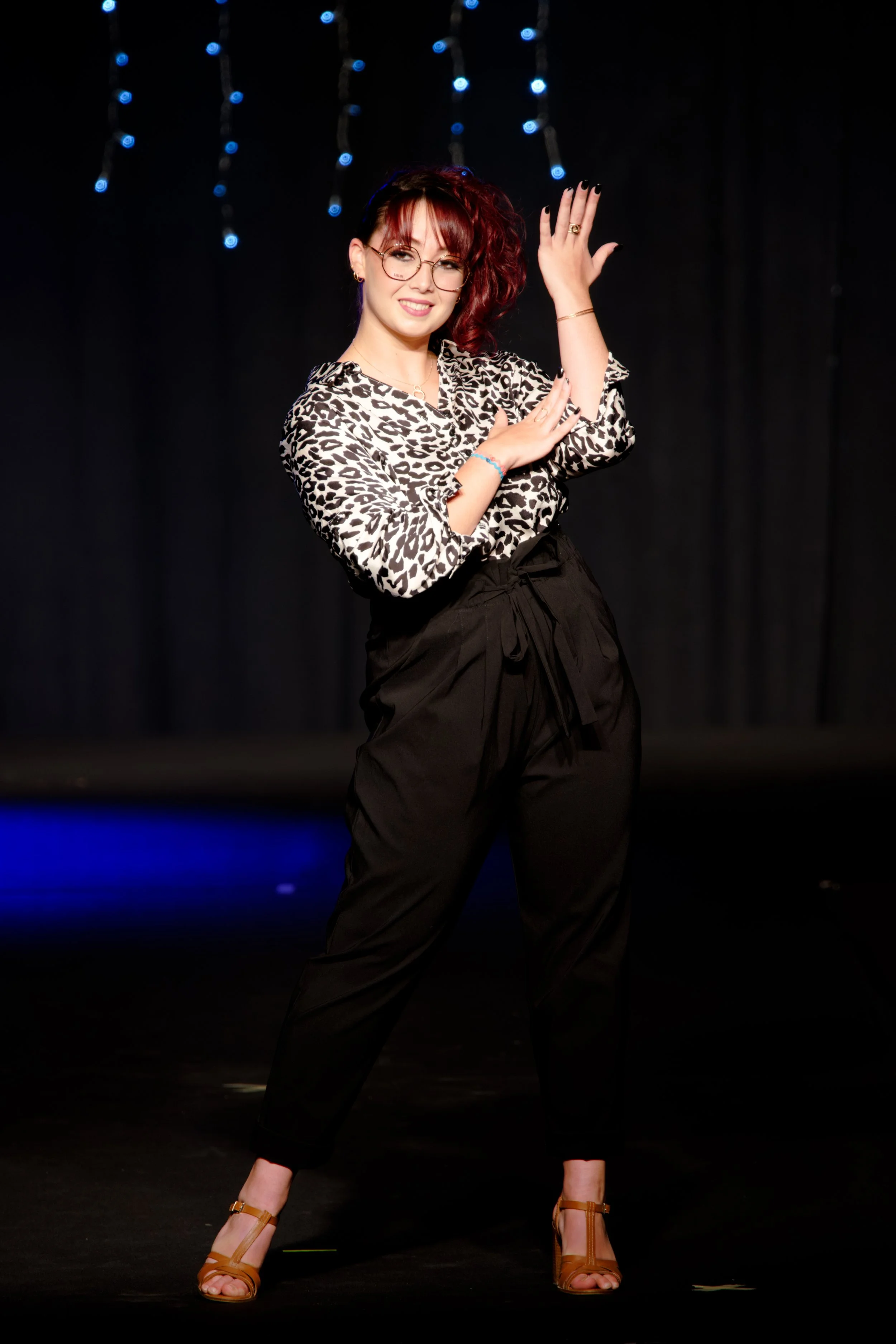Personne aux cheveux rouges, avec des lunettes et un chemisier à imprimé léopard, posant sur une scène faiblement éclairée avec des guirlandes lumineuses.