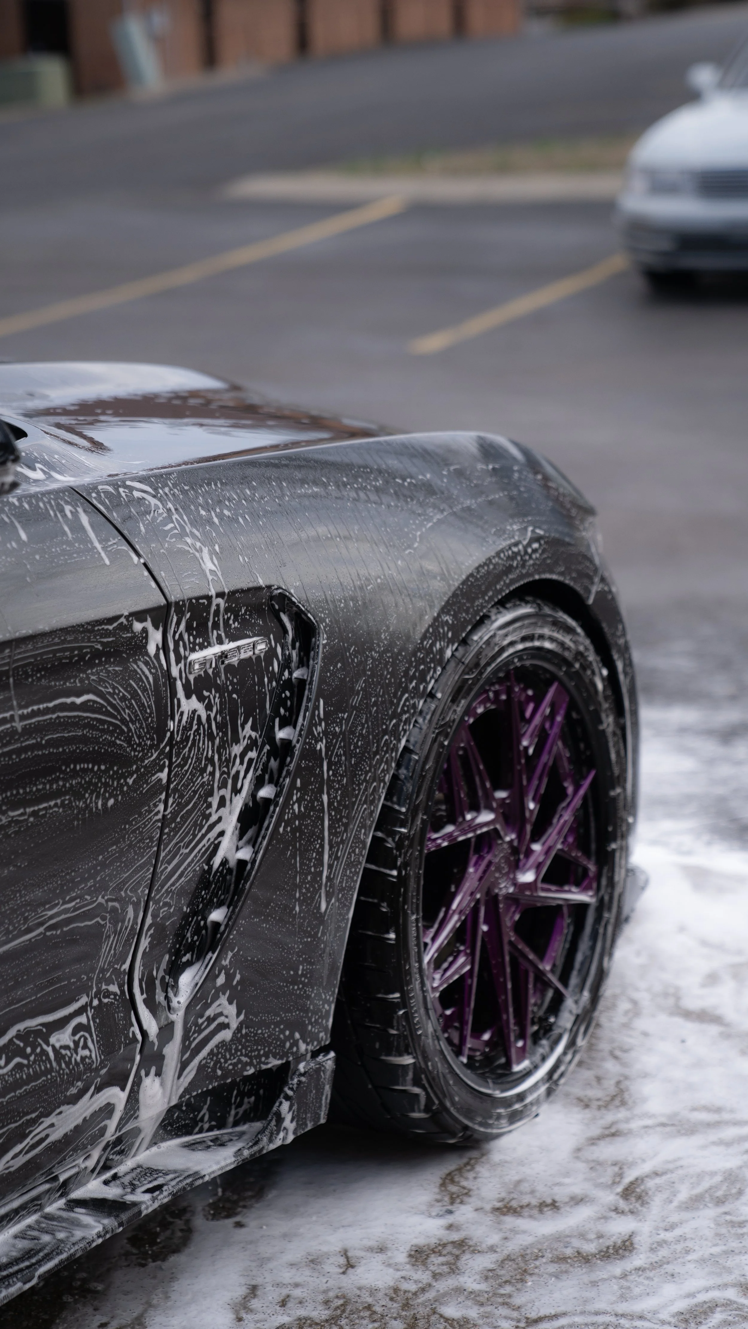Close-up of a black sports car with purple rims being washed, soap suds on the body, in a parking lot with other cars and a brick building in the background.