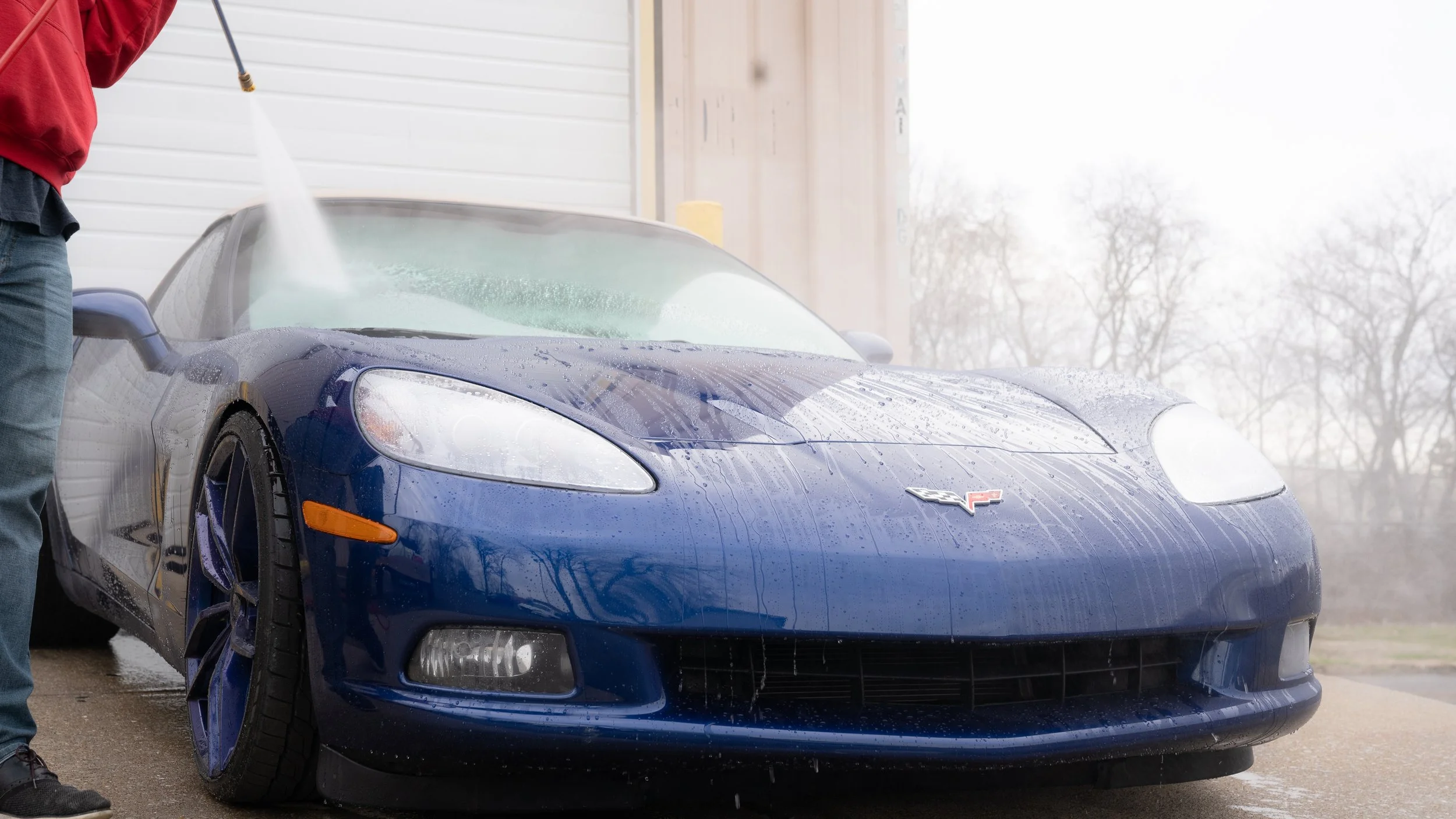 A person washing a blue Chevrolet Corvette with a hose, creating soap suds and water spray, in front of a garage.