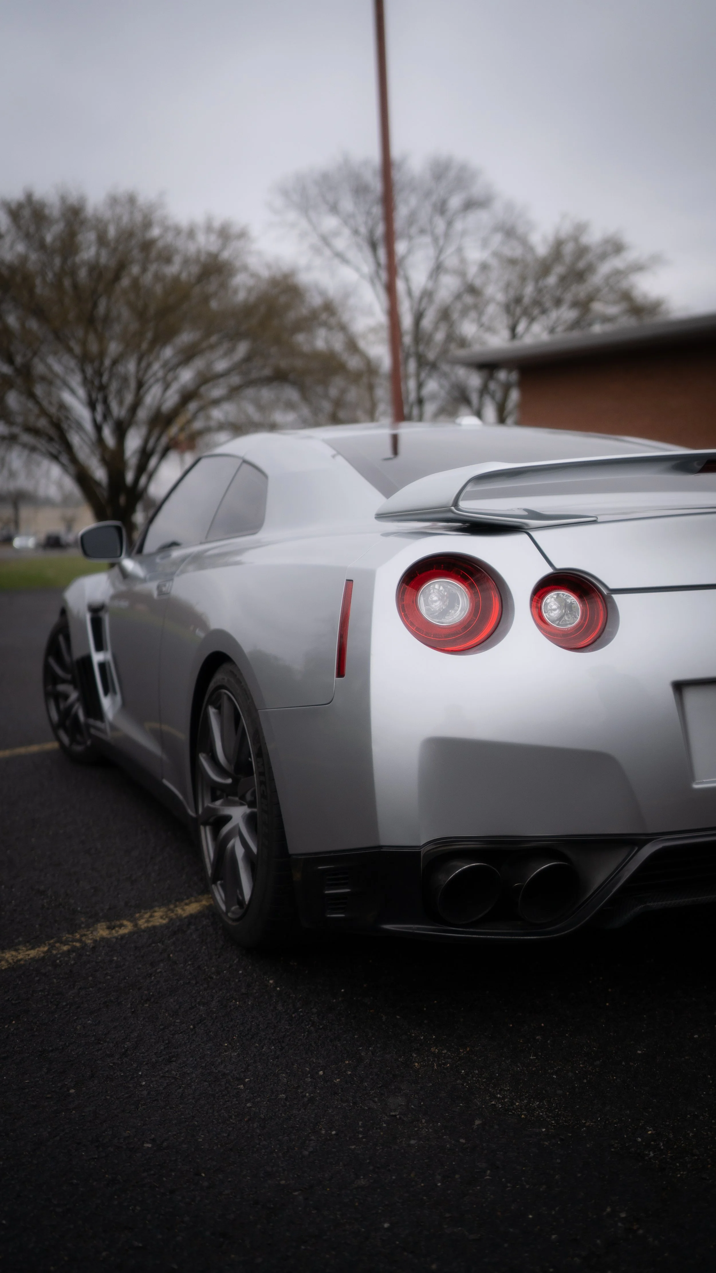 Silver sports car parked on a parking lot with trees and a building in the background under an overcast sky.