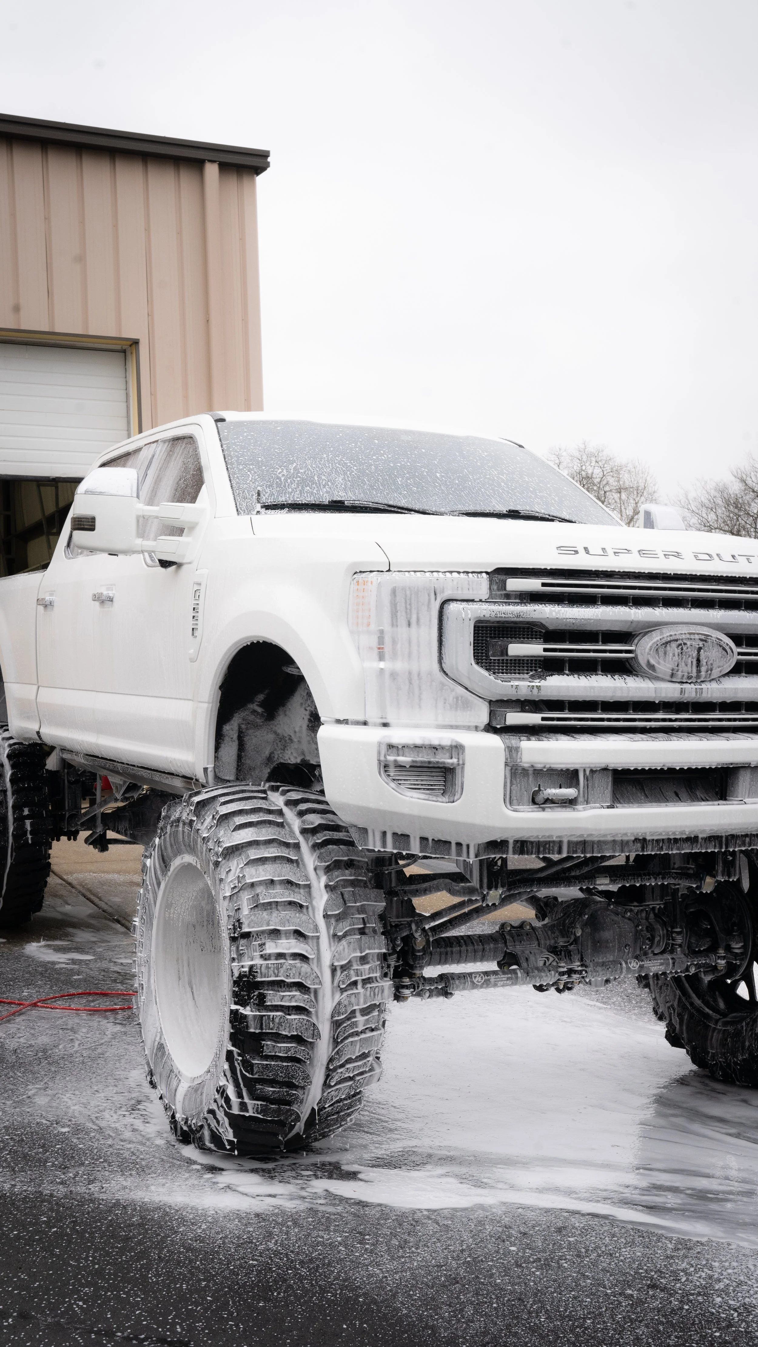 A white Ford SUV with oversized mud tires being washed, covered in soap suds and foam, outside a building with a beige exterior and a garage door.