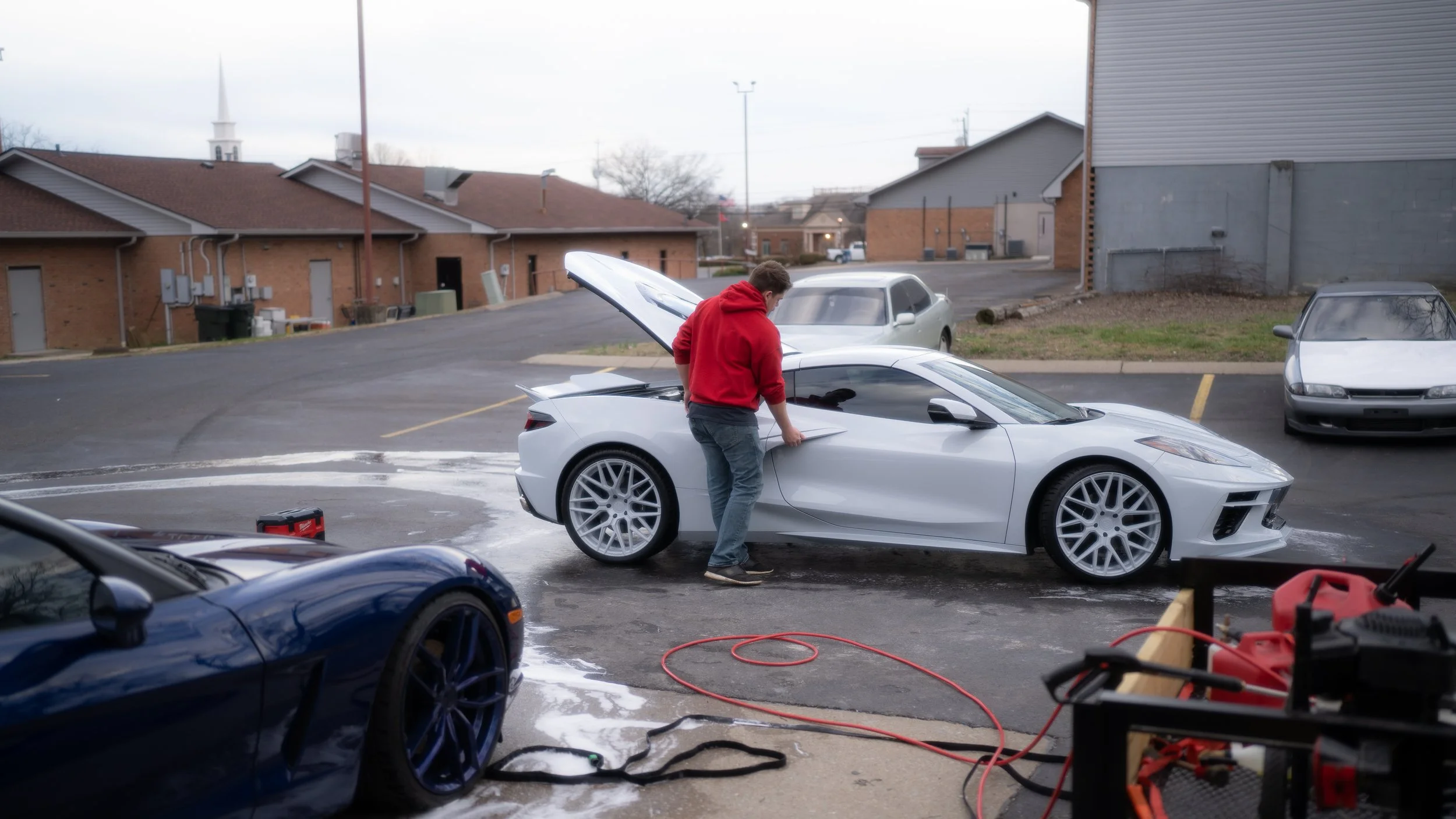 A man in a red hoodie cleaning a white sports car in a parking lot, with open hood and other parked cars around, overcast sky.