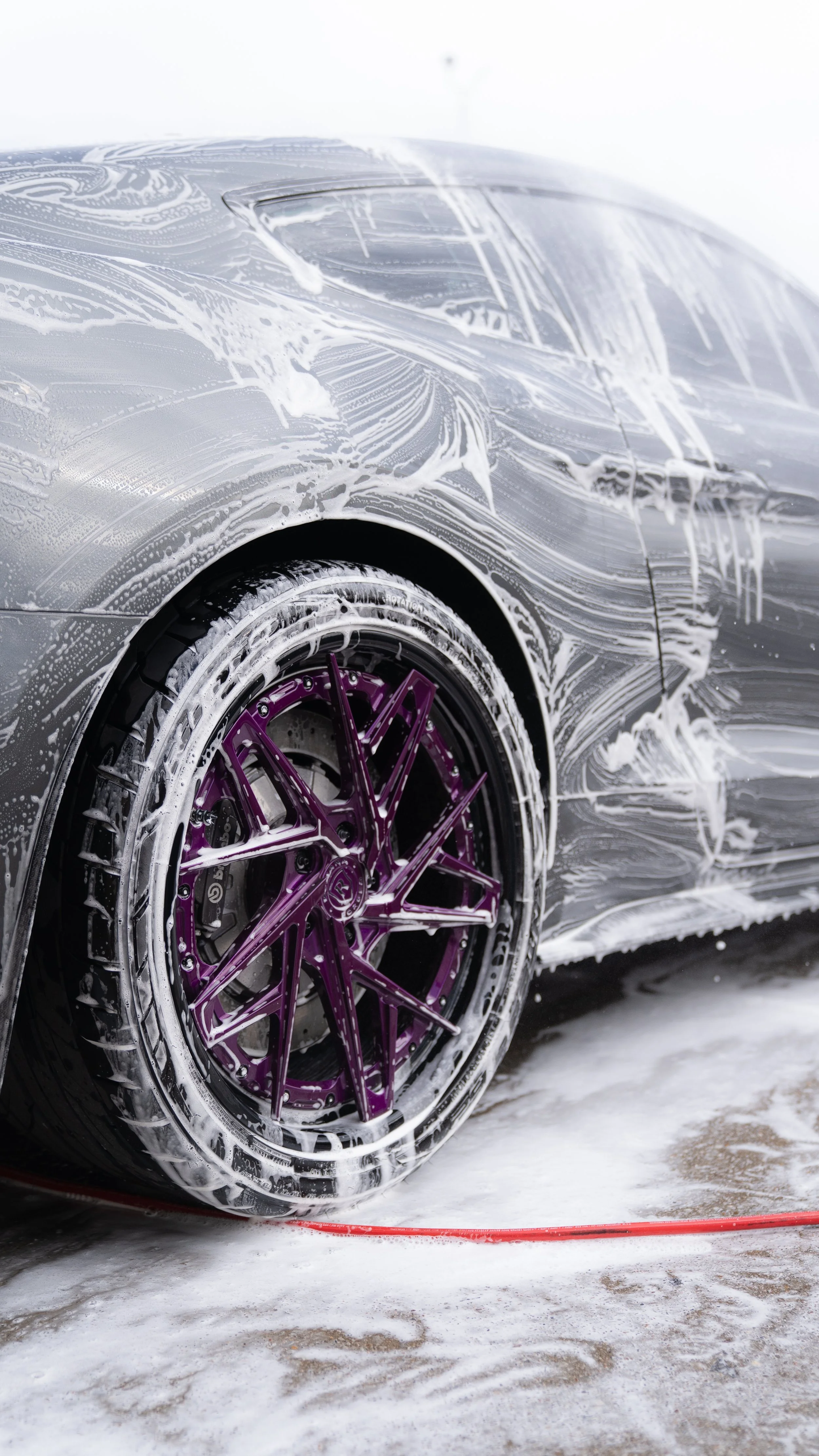 Close-up of a silver car with purple wheels being washed with soap and foam, on a concrete surface.