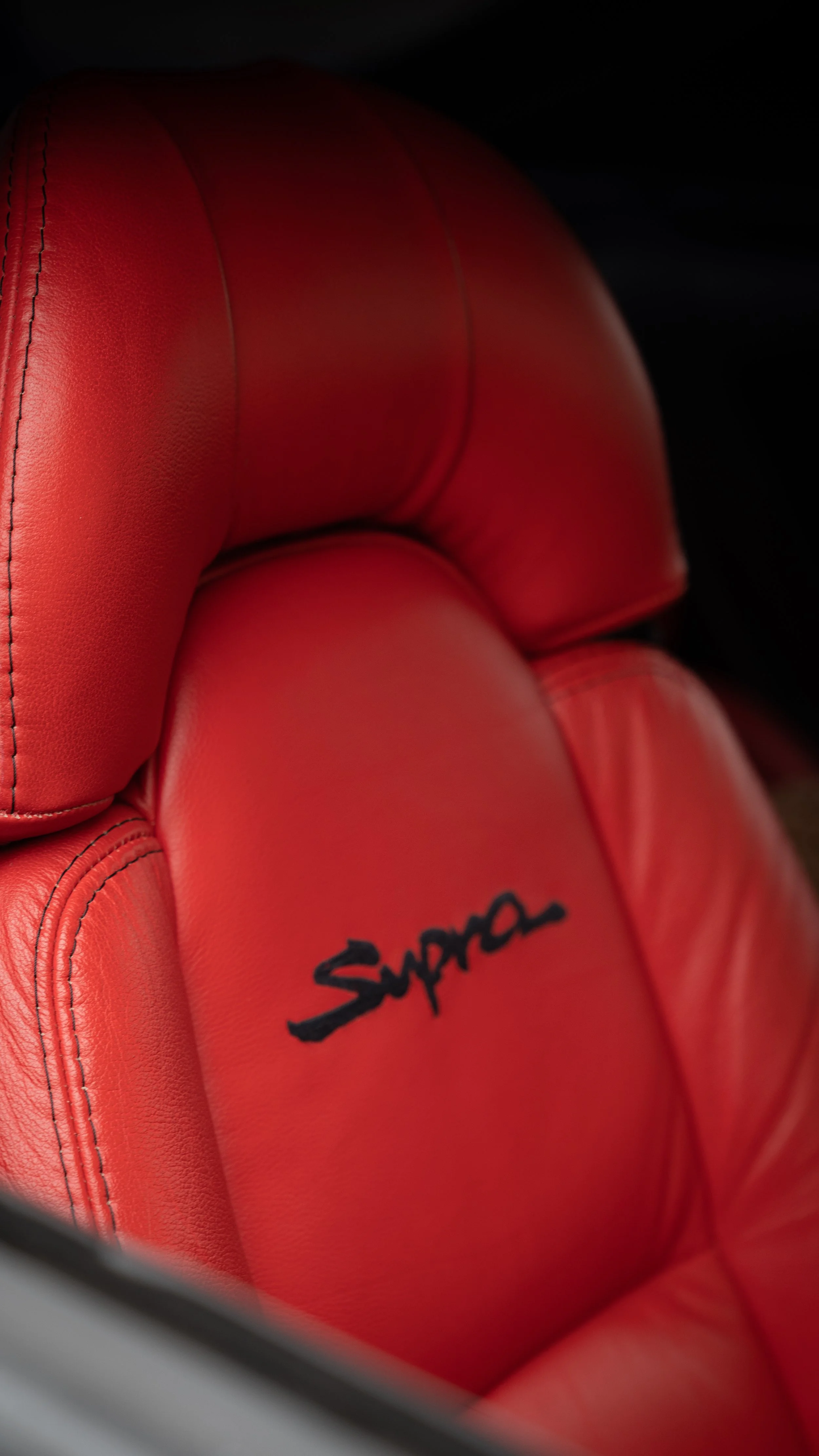 Close-up of a red leather car seat with embroidered black signature that reads 'Supera'.