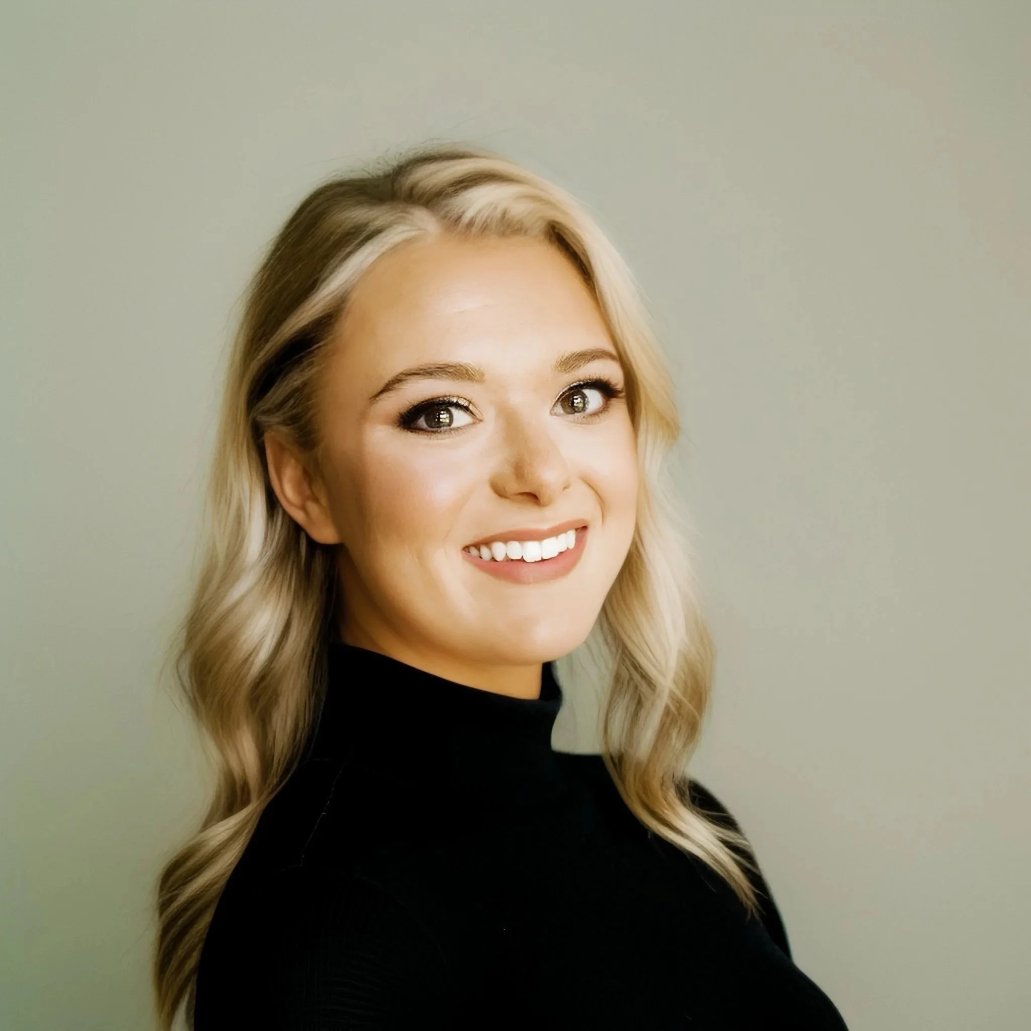 Portrait of a smiling woman with long blonde hair wearing a black turtleneck sweater, against a plain green background.