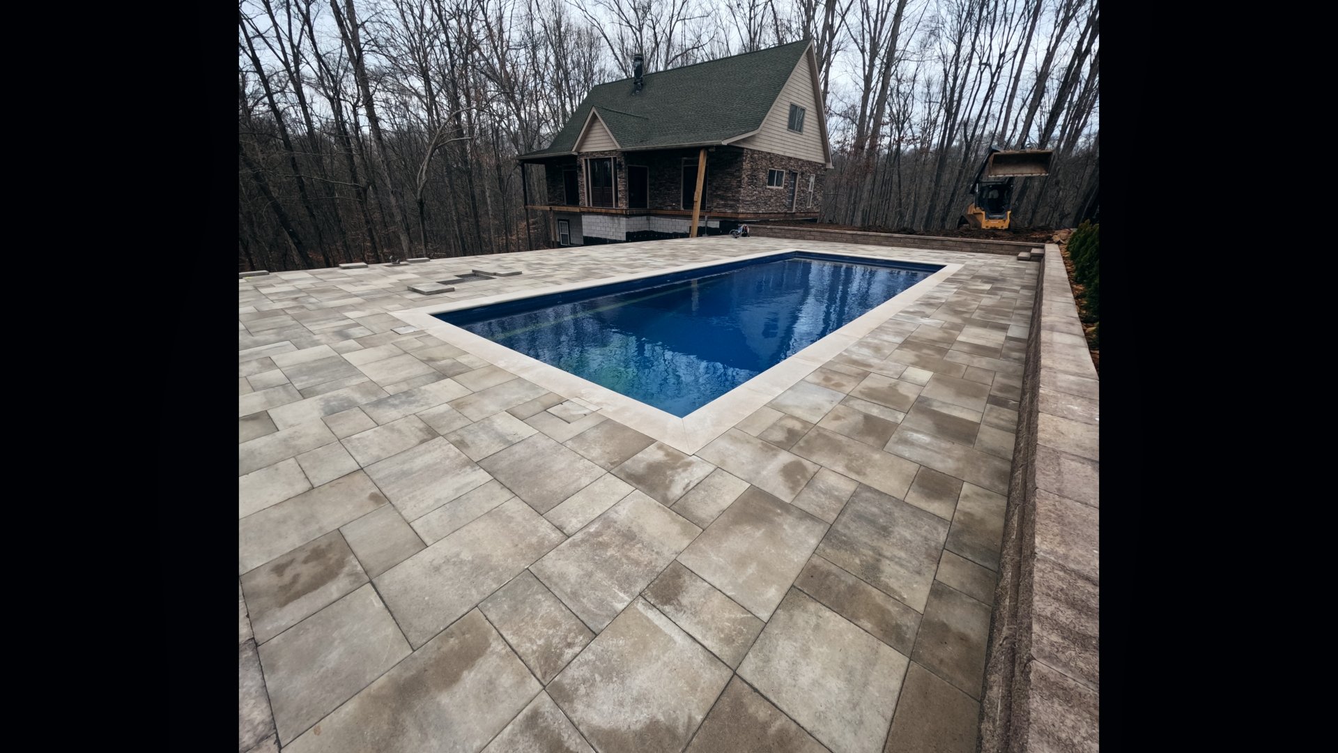 House with swimming pool and stone patio surrounded by bare trees in a forested area.