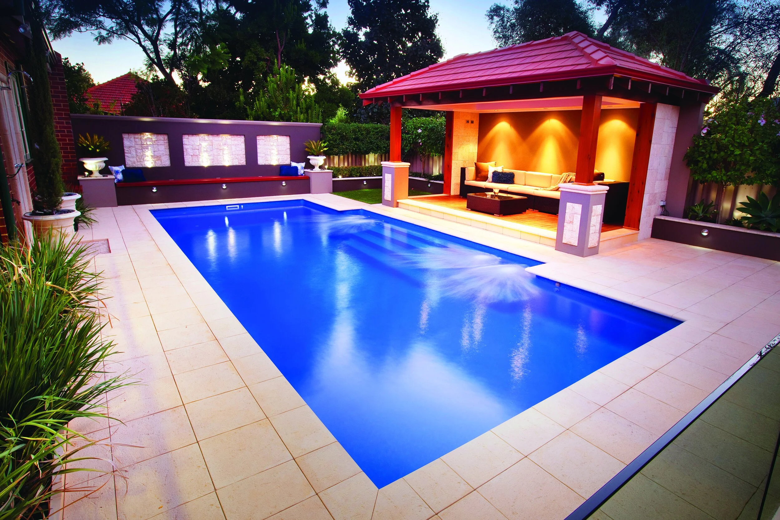 Modern backyard with illuminated swimming pool, tiled patio, seating area under a gazebo, plants, and ambient lighting at dusk.