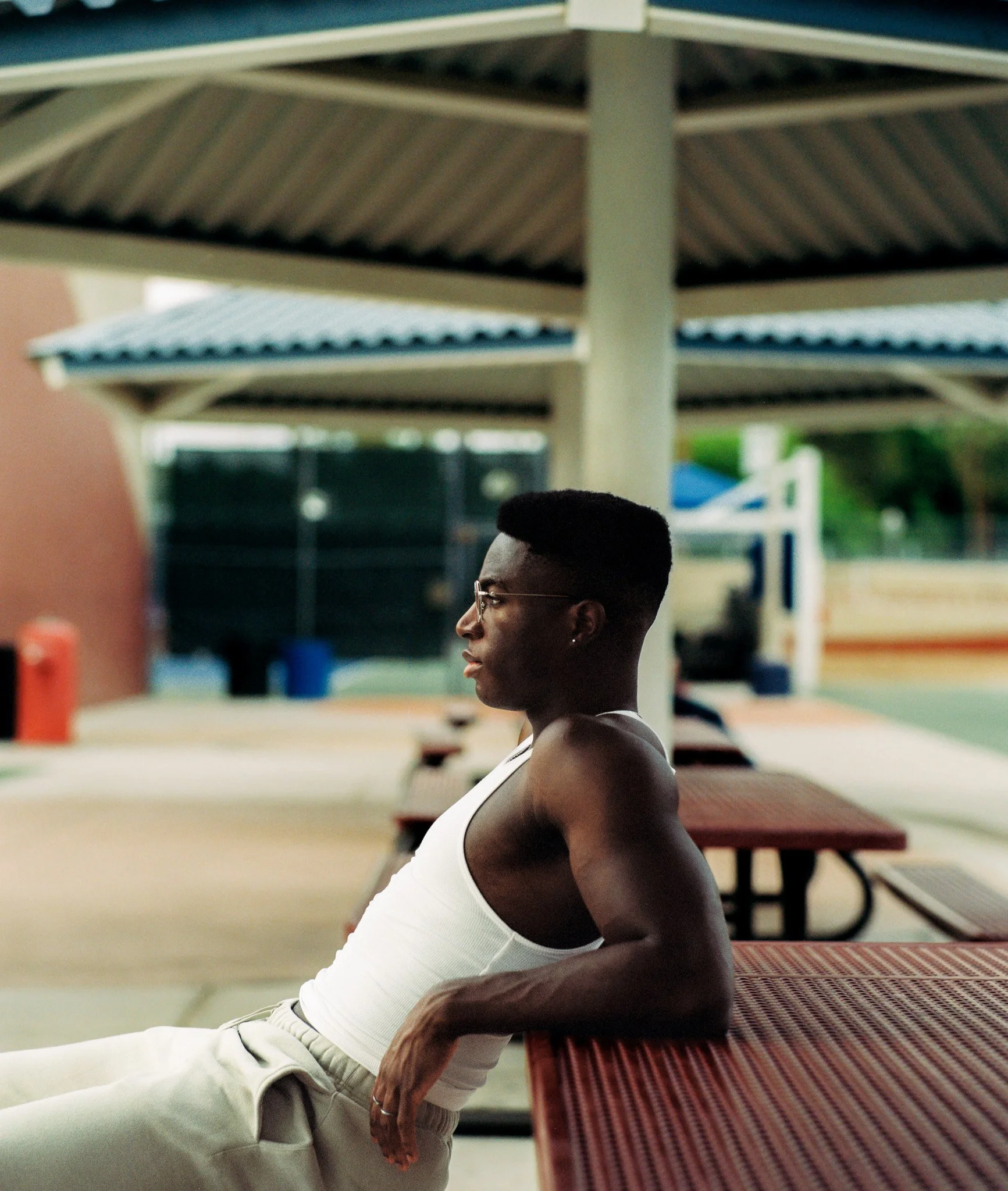 A person with short hair and glasses, wearing a white tank top and beige pants, sitting at an outdoor location with a stage or shelter with a striped roof in the background.