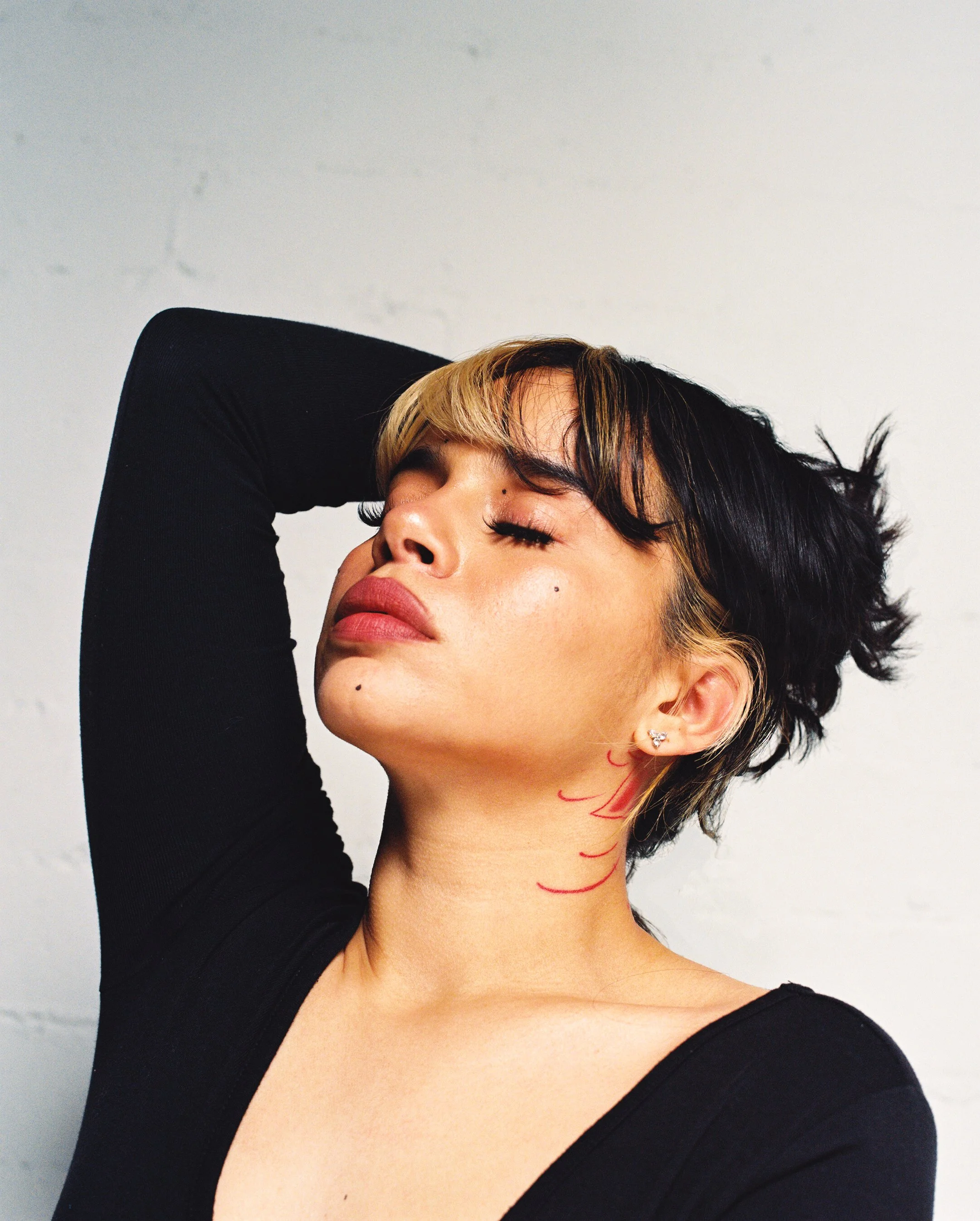 A woman with short black and blonde hair, eyes closed, wearing earrings, with red marks on her neck, against a plain light-colored background.