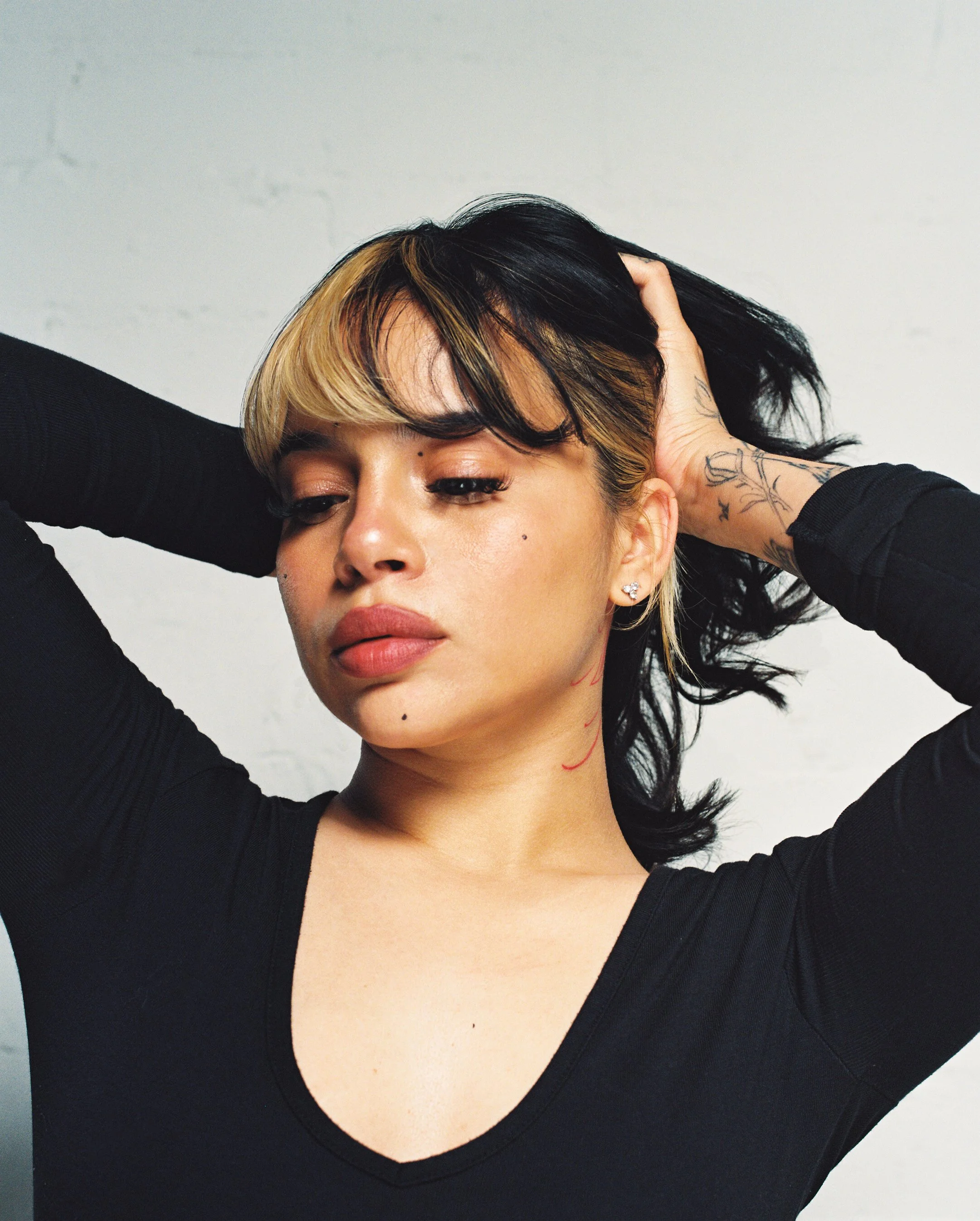 Close-up of a woman with short black hair with blonde highlights, wearing a black top, touching her head with both hands, and looking down slightly against a light background.
