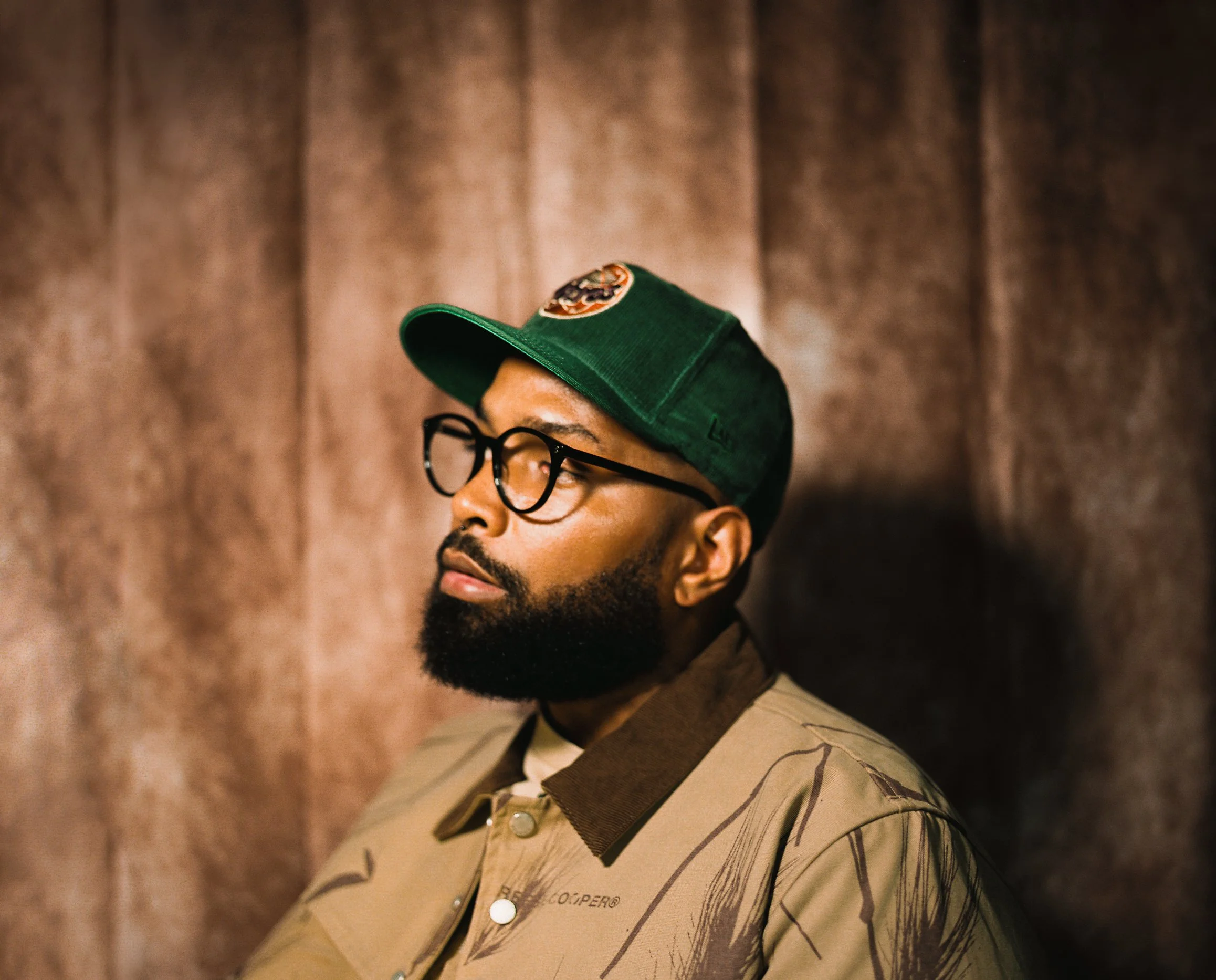 A young man with glasses and facial hair wearing a green baseball cap with a logo and a beige button-up shirt with a leaf pattern. The background is a brown textured wall.