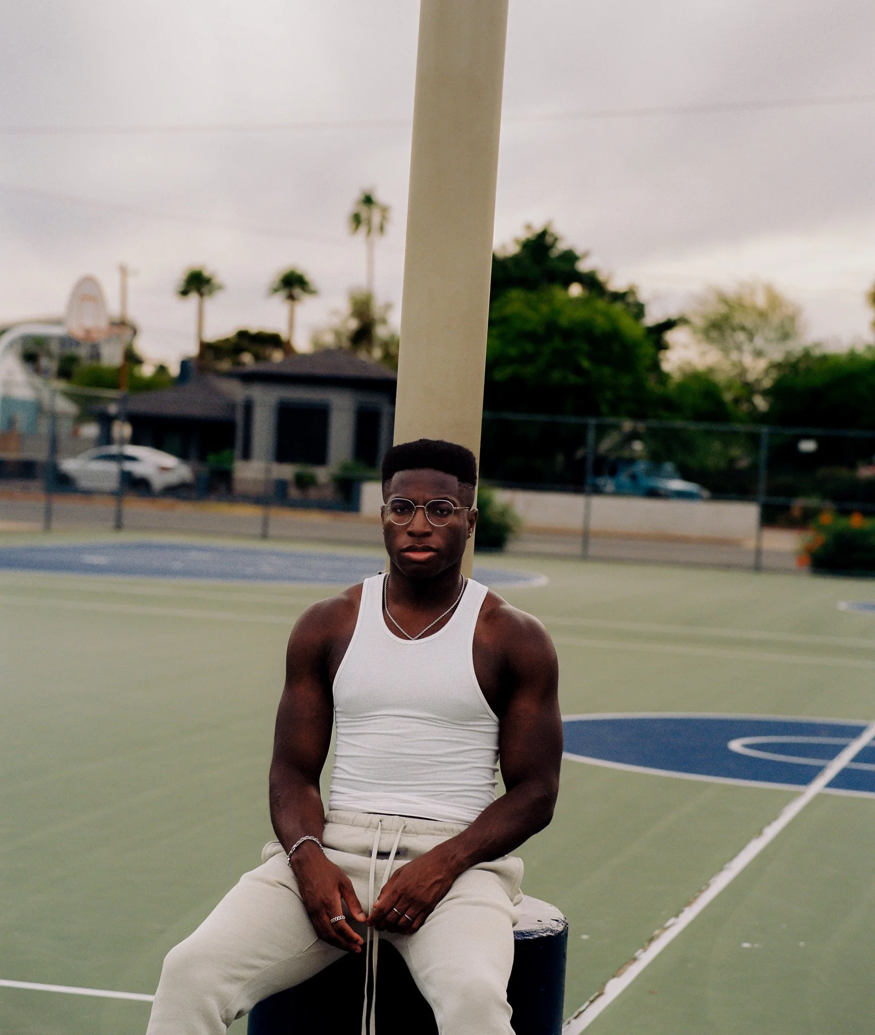 A young man with dark skin, wearing a white tank top and beige pants, sitting on a black stool at an outdoor basketball court. The background features palm trees, residential houses, a white car, and a basketball hoop under a cloudy sky.
