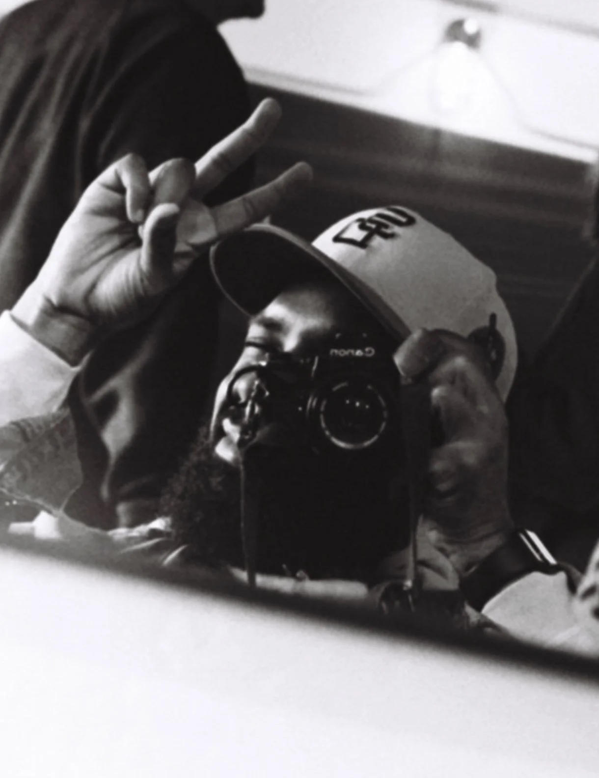Black and white photo of a person taking a selfie mirror with a camera. The person is wearing a baseball cap, glasses, and a hoodie, making a peace sign with one hand.