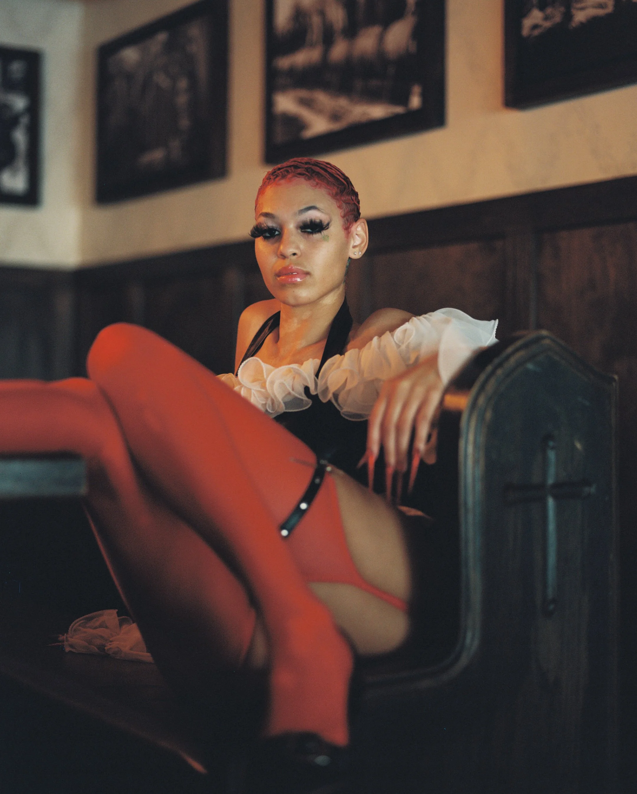 A woman with red braided hair and makeup sitting on a chair, wearing a white ruffled off-shoulder top and black straps, in a room with framed pictures on the wall.