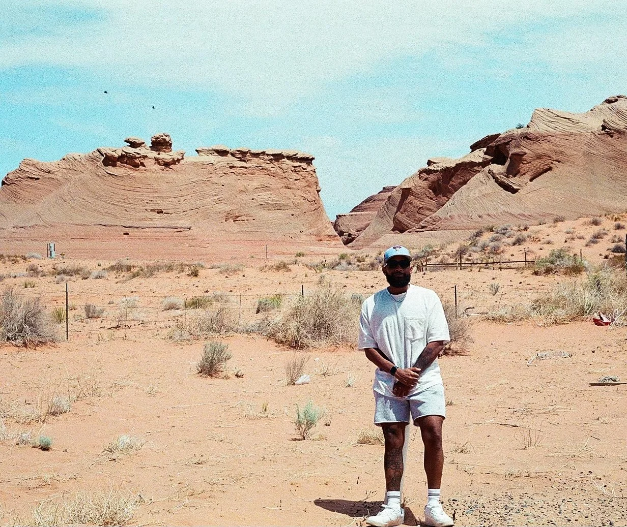 A man in white casual clothes, sunglasses, and a cap stands in a desert with rocky formations and sparse vegetation under a clear blue sky.