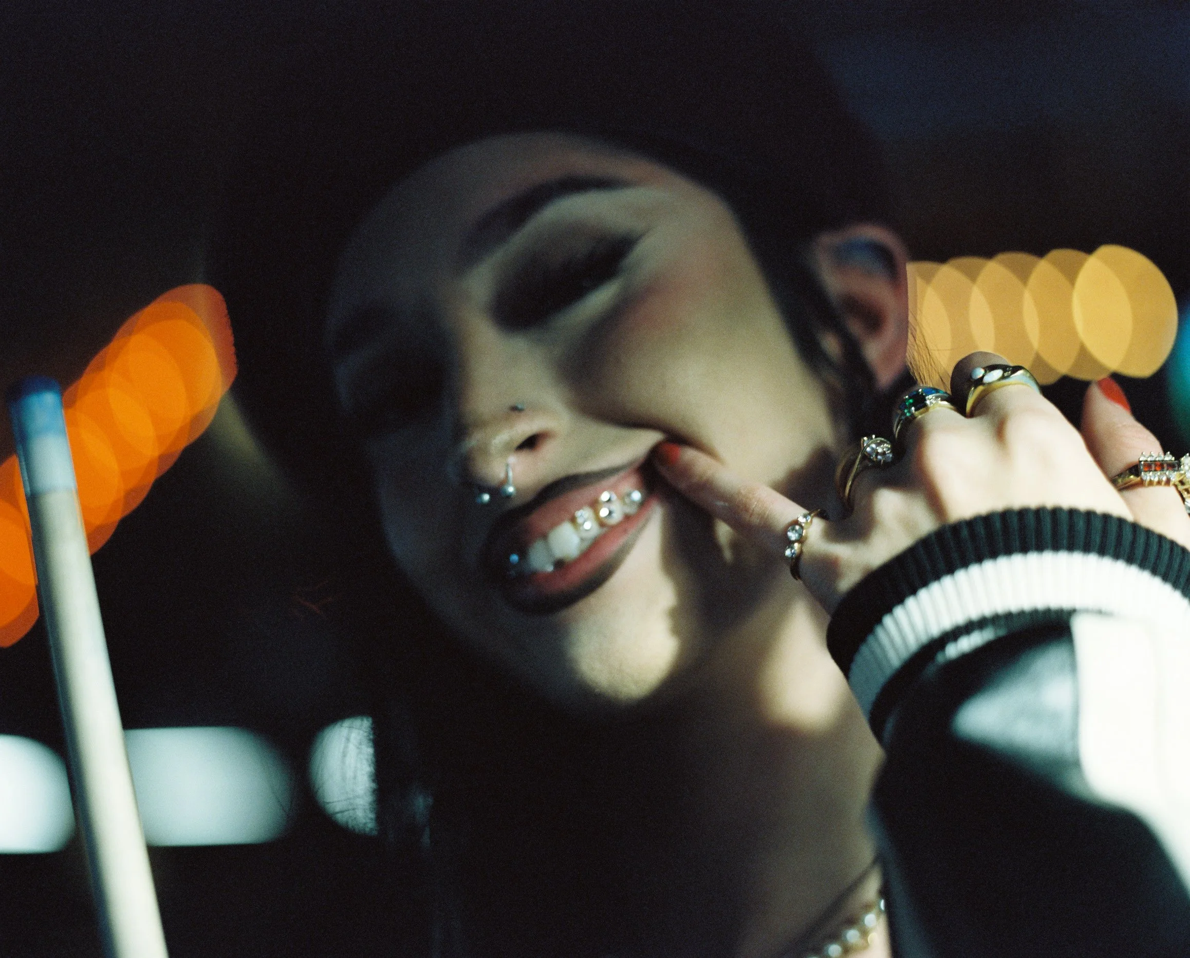 Close-up of a woman's face with makeup, jewelry, and a hand near her lips, viewed through circular objects or reflections, with colorful blurred background lights.
