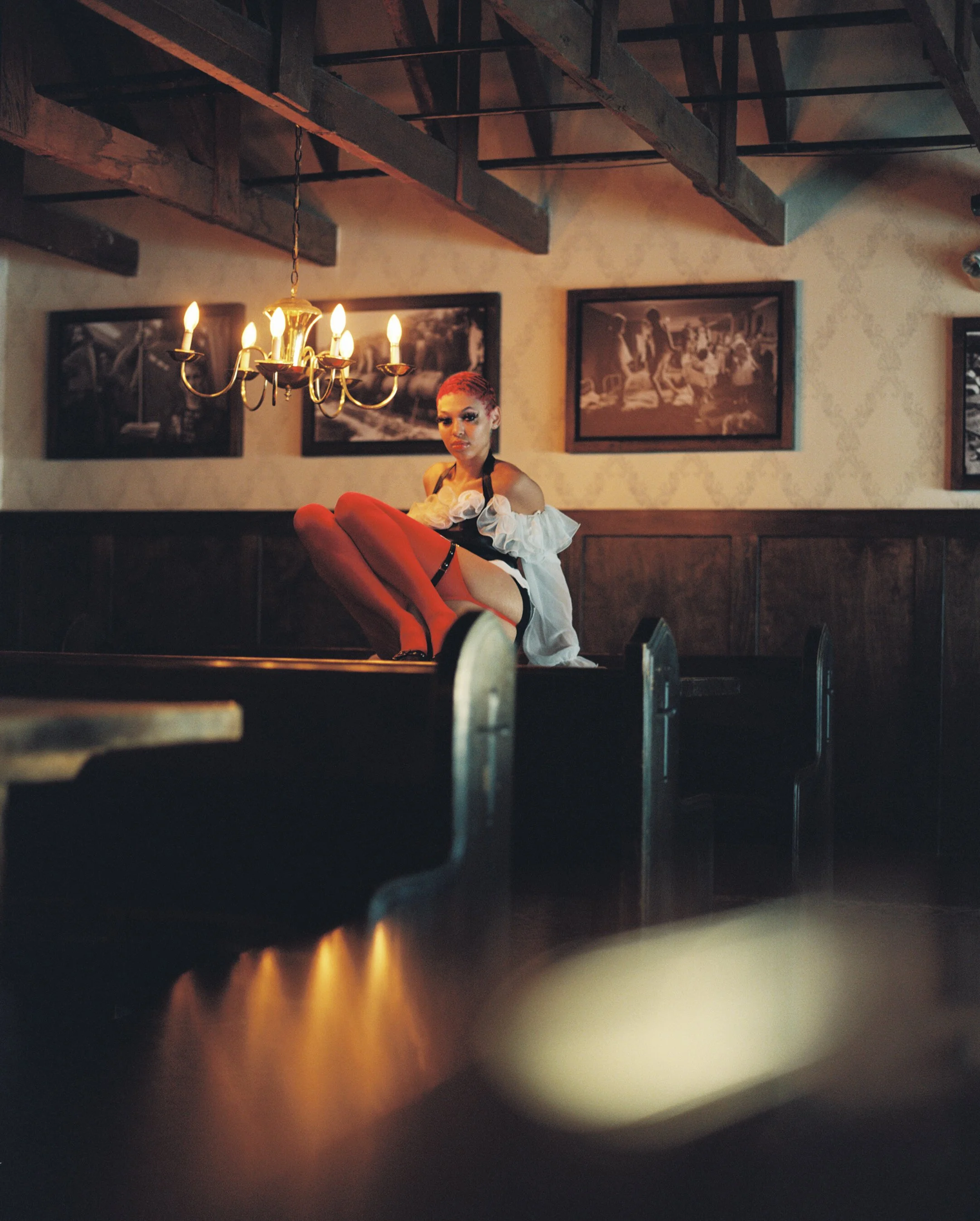 Dimly lit scene inside a restaurant with a chandelier hanging from the ceiling, featuring a woman sitting at a table. The woman has bright red hair and is wearing a white off-shoulder top and red boots. The walls are decorated with framed black-and-w