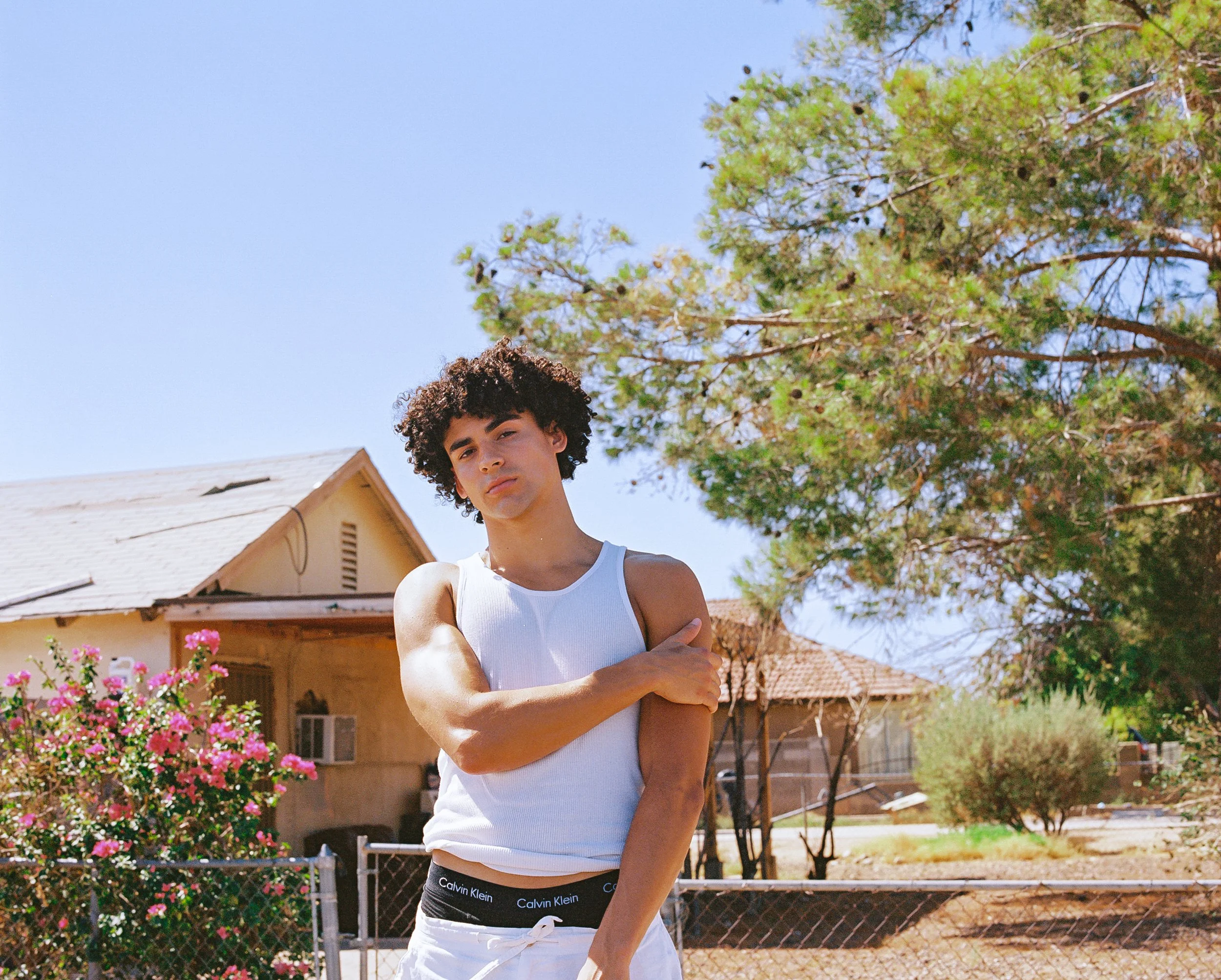 Young man with curly dark hair wearing a white tank top and black Calvin Klein underwear standing outside in front of a house with trees and pink flowers.