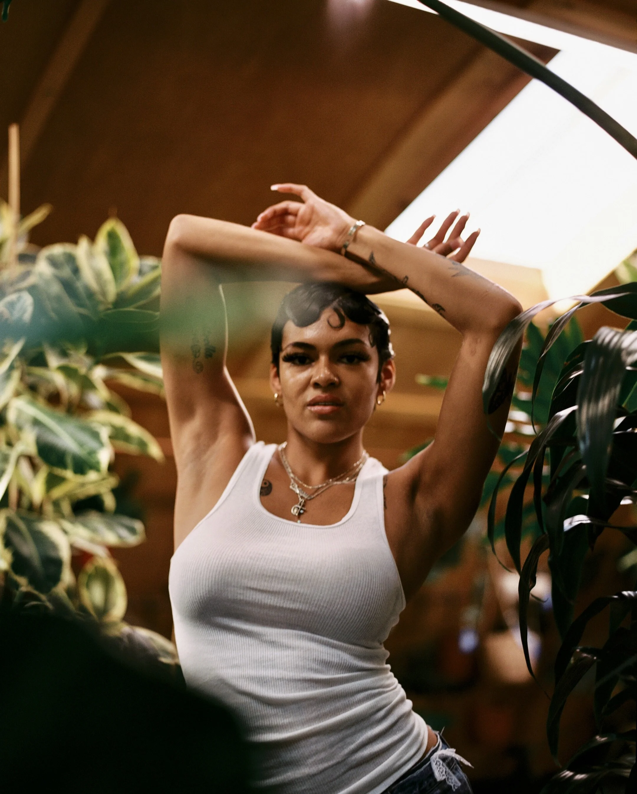 A woman with dark hair and tattoos on her arms is gazing directly into the camera, partially covered by plants, and standing in a warm, indoor setting with wooden walls and a large white ceiling.