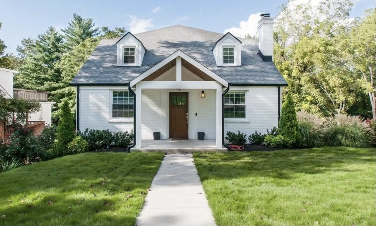 Front view of a white house with a gray roof, small front porch, and a concrete pathway leading to the entrance, surrounded by green lawn and trees.