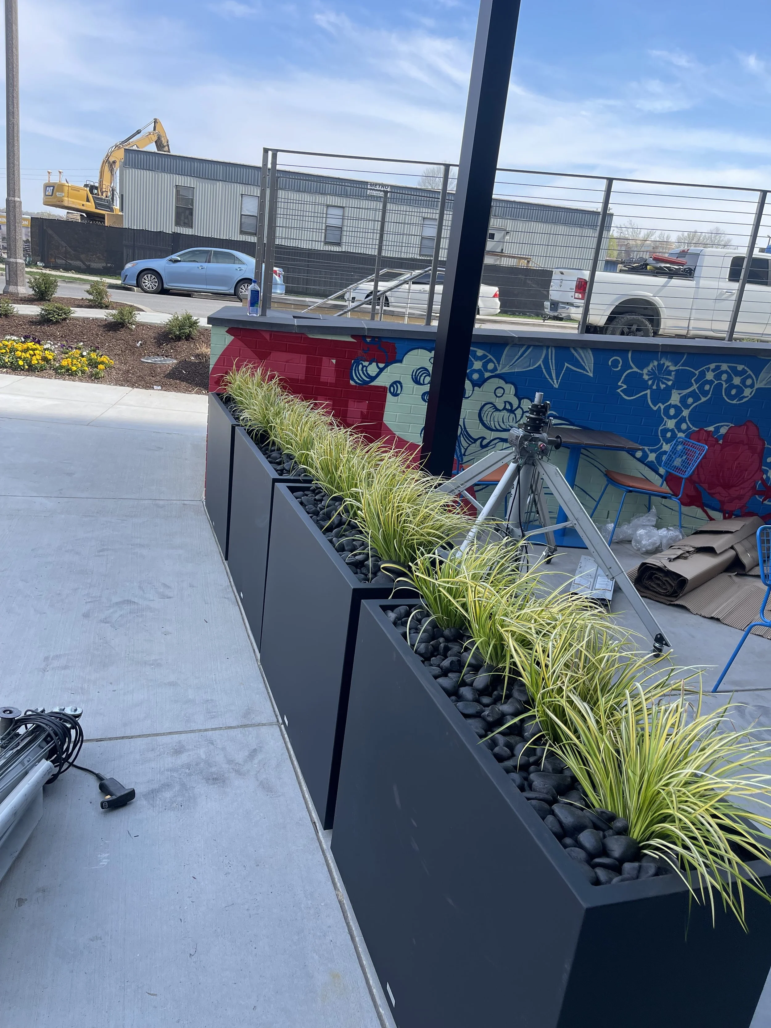 Long black planter box filled with ornamental grass and black stones, set on a sidewalk outside a building with a colorful mural.