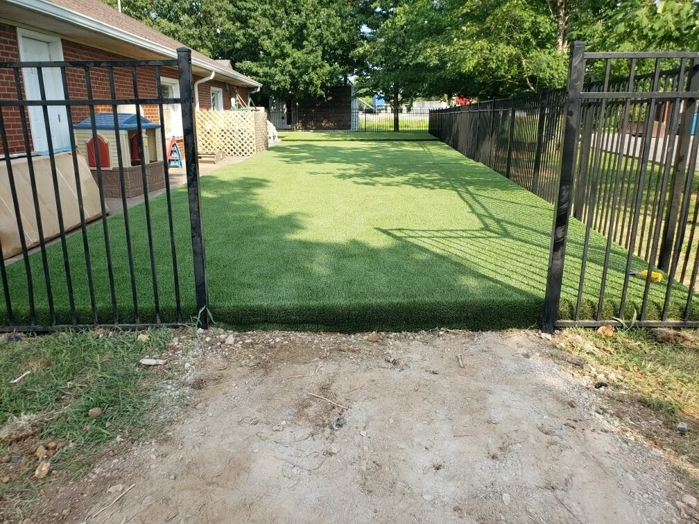 A backyard with a black metal fence enclosing an artificially turfed lawn area next to a brick house. There are trees in the background, with some shadows cast on the grass.