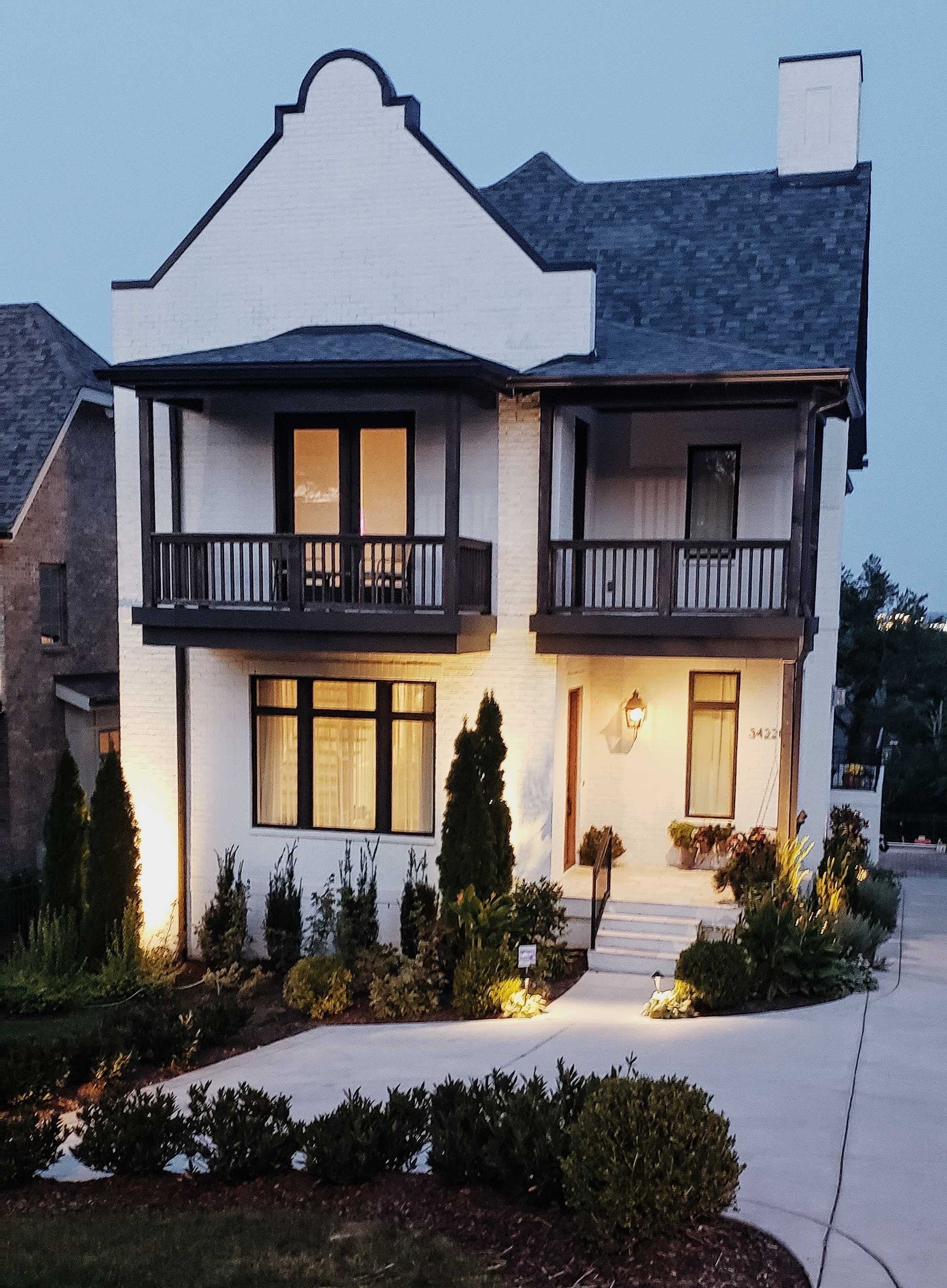 A two-story modern house with a white exterior, black balcony railings, and a dark roof. The house has large front windows, a front porch with steps, and is illuminated by exterior lighting. There are landscaped bushes and plants in the front yard.