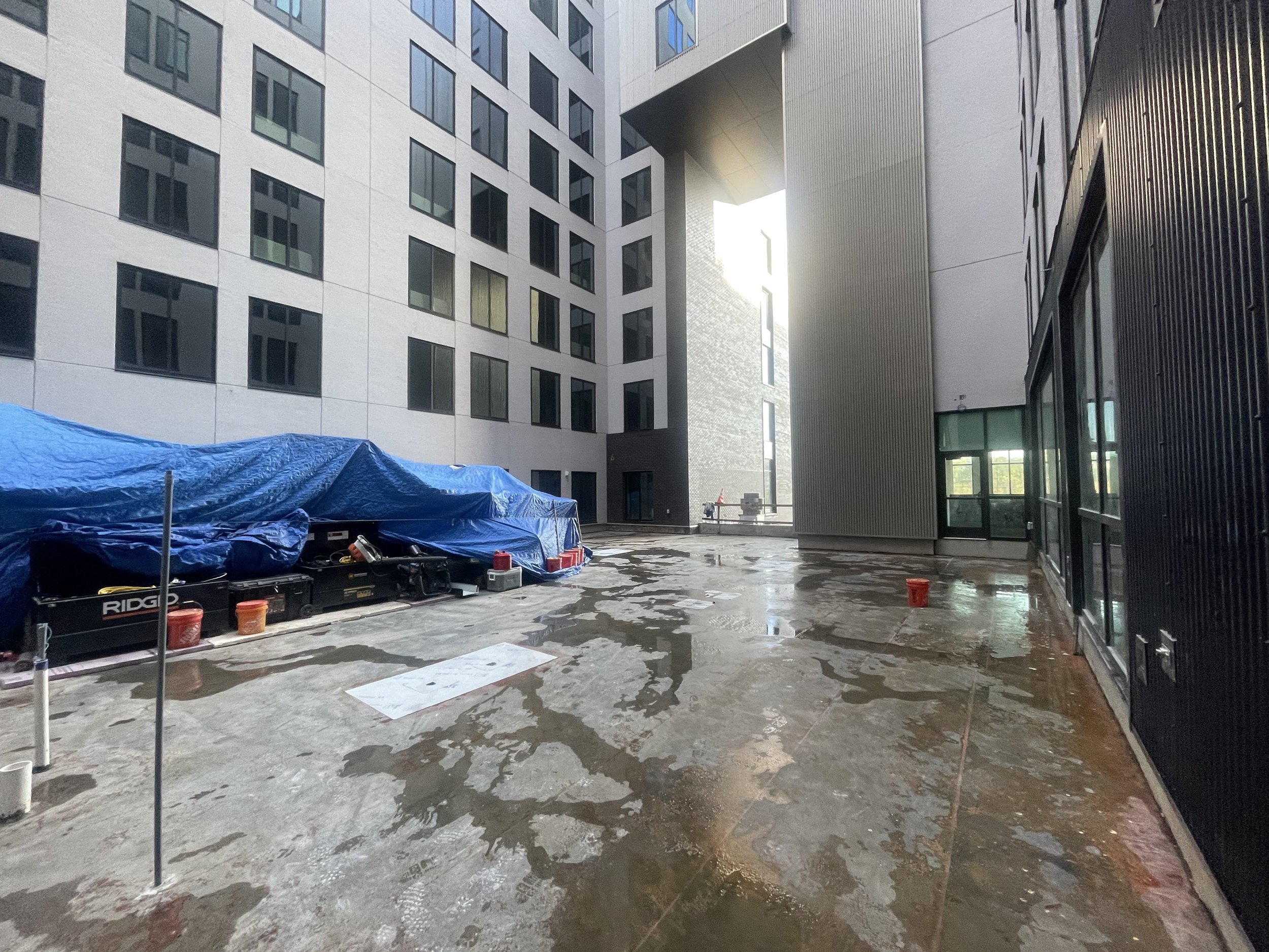 Construction site on a wet concrete floor surrounded by tall modern buildings, with a blue tarp covering equipment and red buckets scattered around.