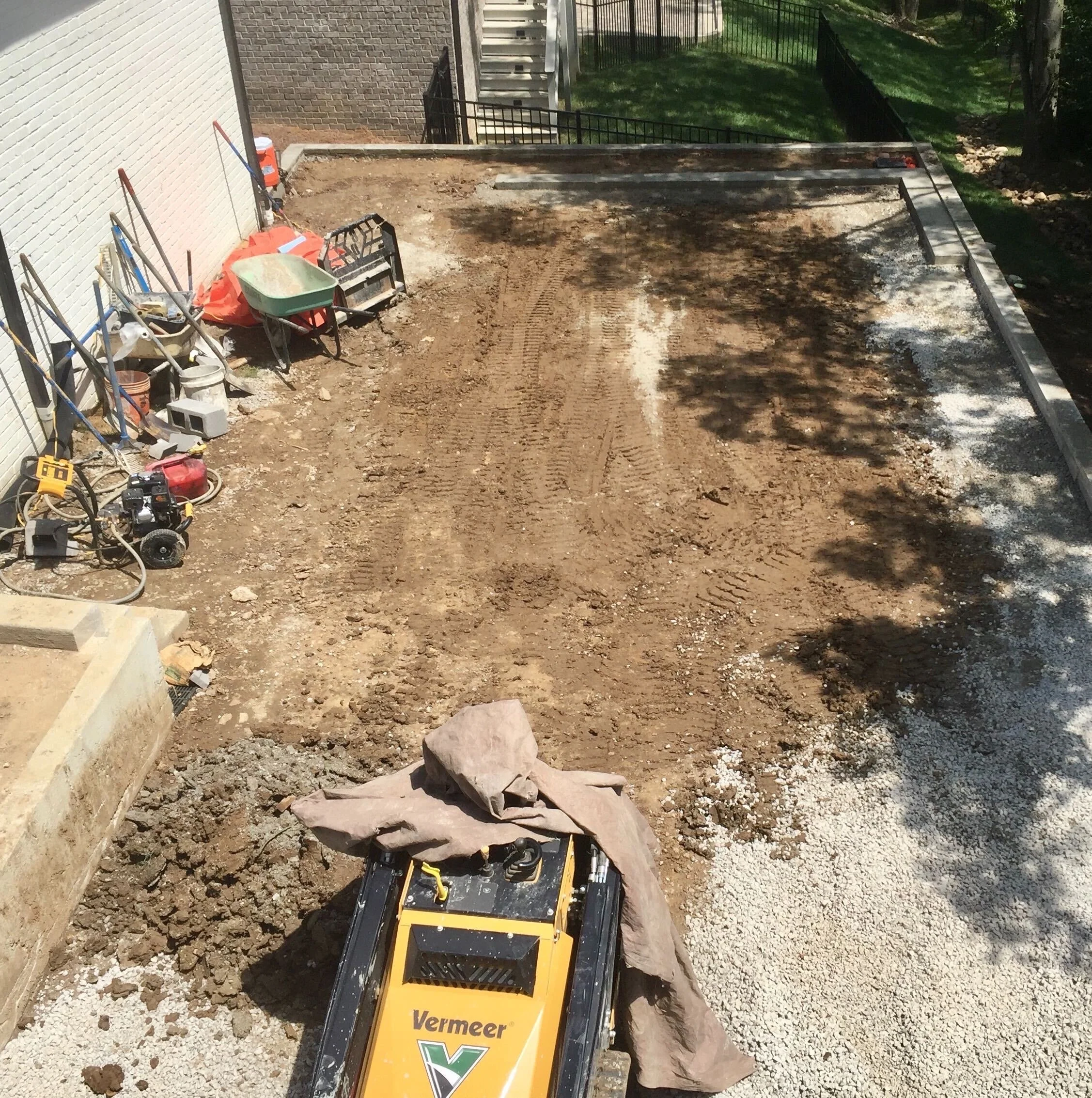 A backyard under construction with a dirt surface, tools, and equipment like a wheelbarrow, shovels, and a small compact excavator.