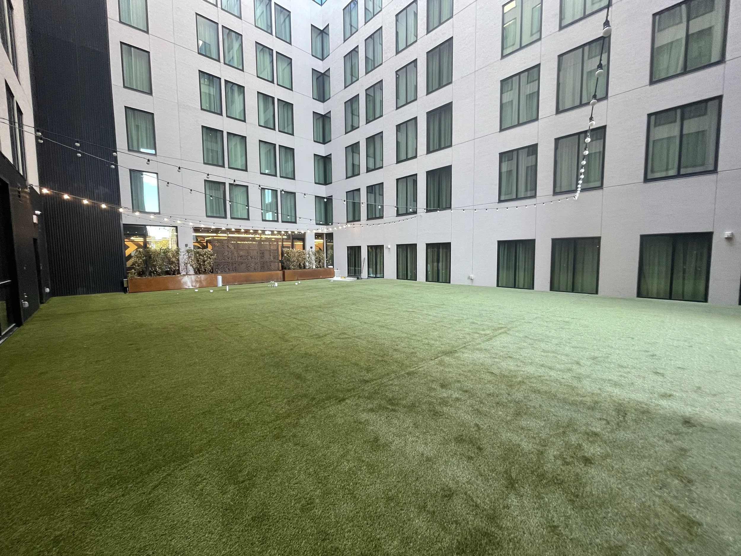 Outdoor courtyard with artificial grass, string lights, and surrounding modern multi-story buildings with numerous windows.