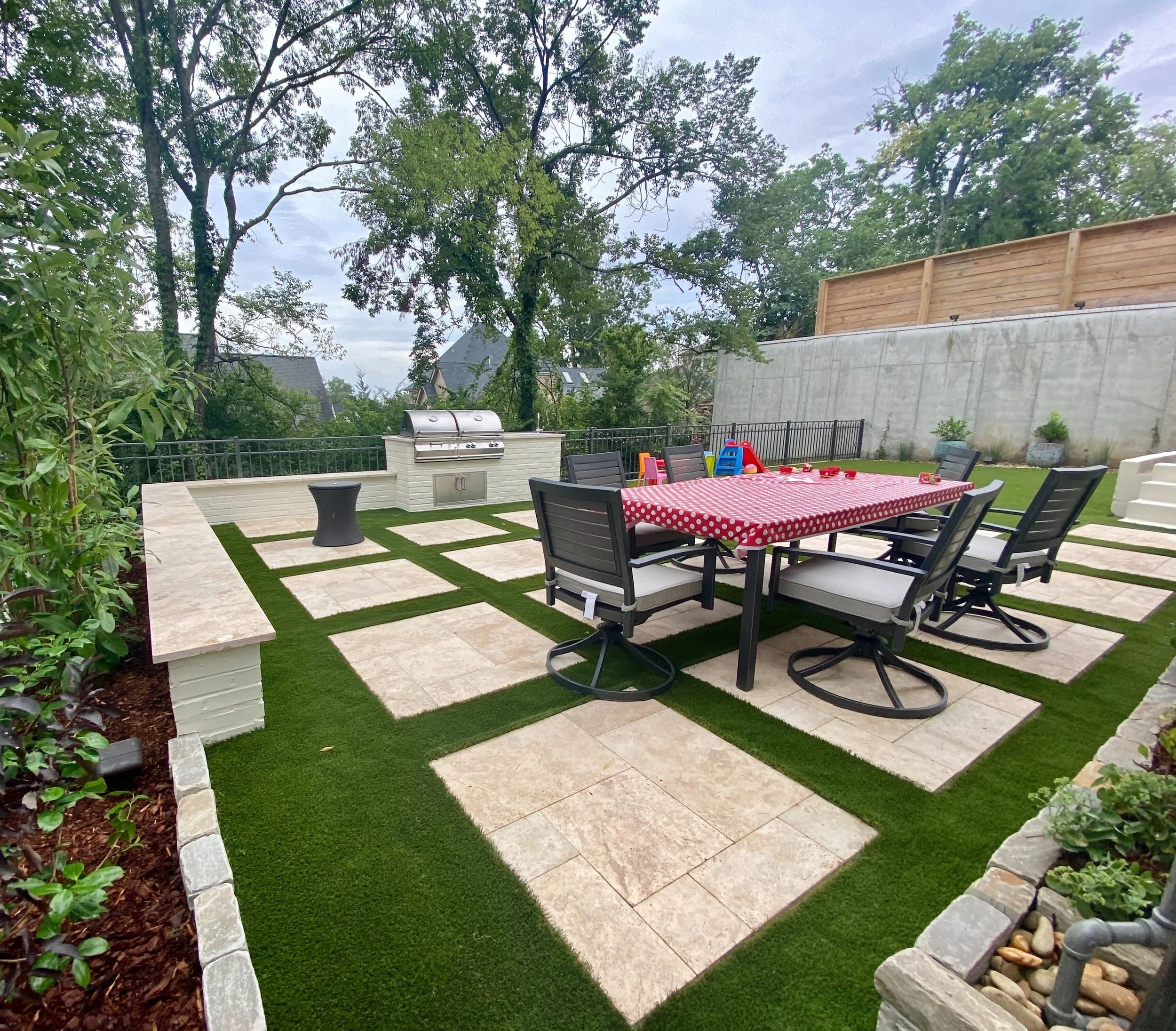 A backyard patio with a dining table covered by a red polka-dotted tablecloth, surrounded by six chairs with white cushions. The area features a tiled and artificial grass patterned flooring, a built-in outdoor grill, and a small side table. There ar