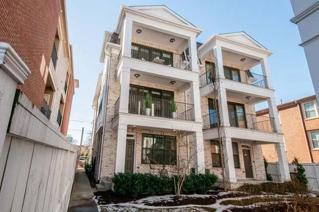 A three-story residential building with white exterior walls, balconies with black railings, and large windows, situated between other buildings, with small bushes and patches of snow on the ground.
