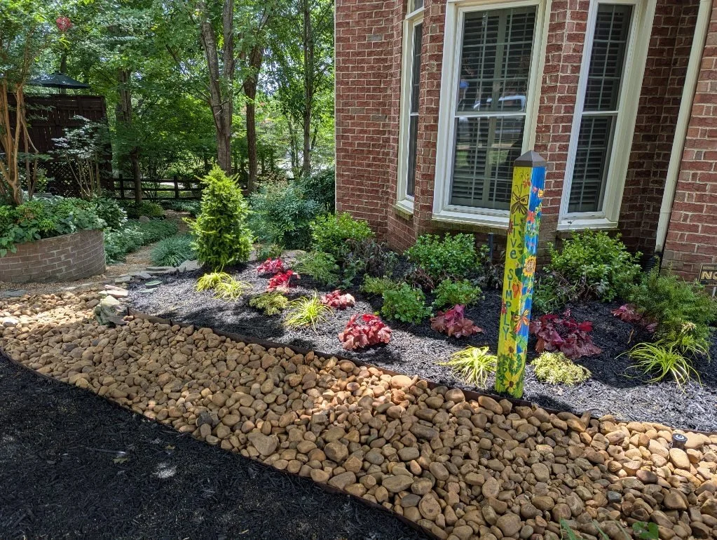 A landscaped garden with a mulch bed containing small green plants and red-leafed plants next to a brick house with white window shutters; a decorative painted wooden sign with flowers and the words 'Love, Simply' stands among the plants; smooth tan 