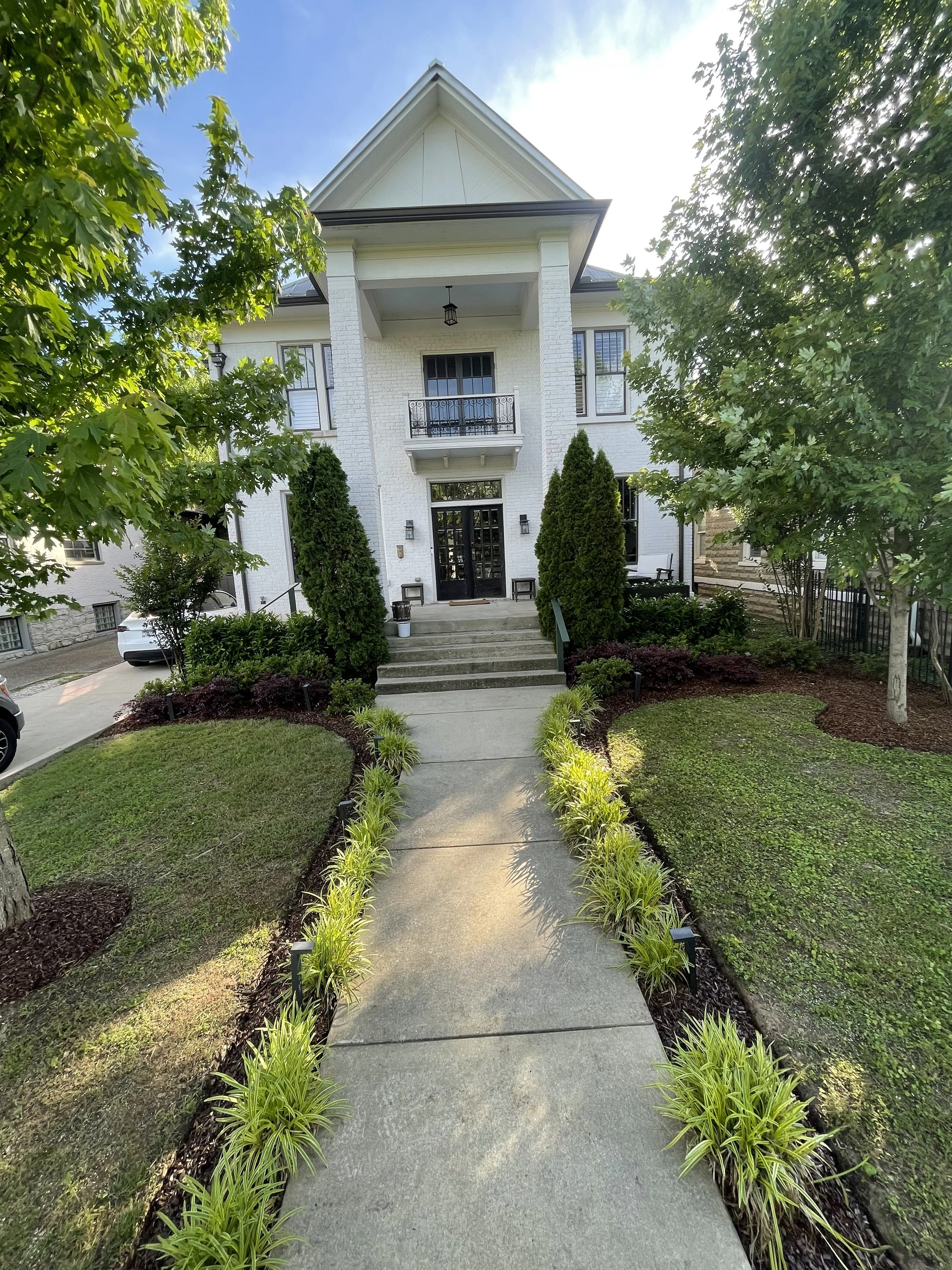 Front view of a white house with front steps, surrounded by greenery, trees, and a landscaped yard on a sunny day.