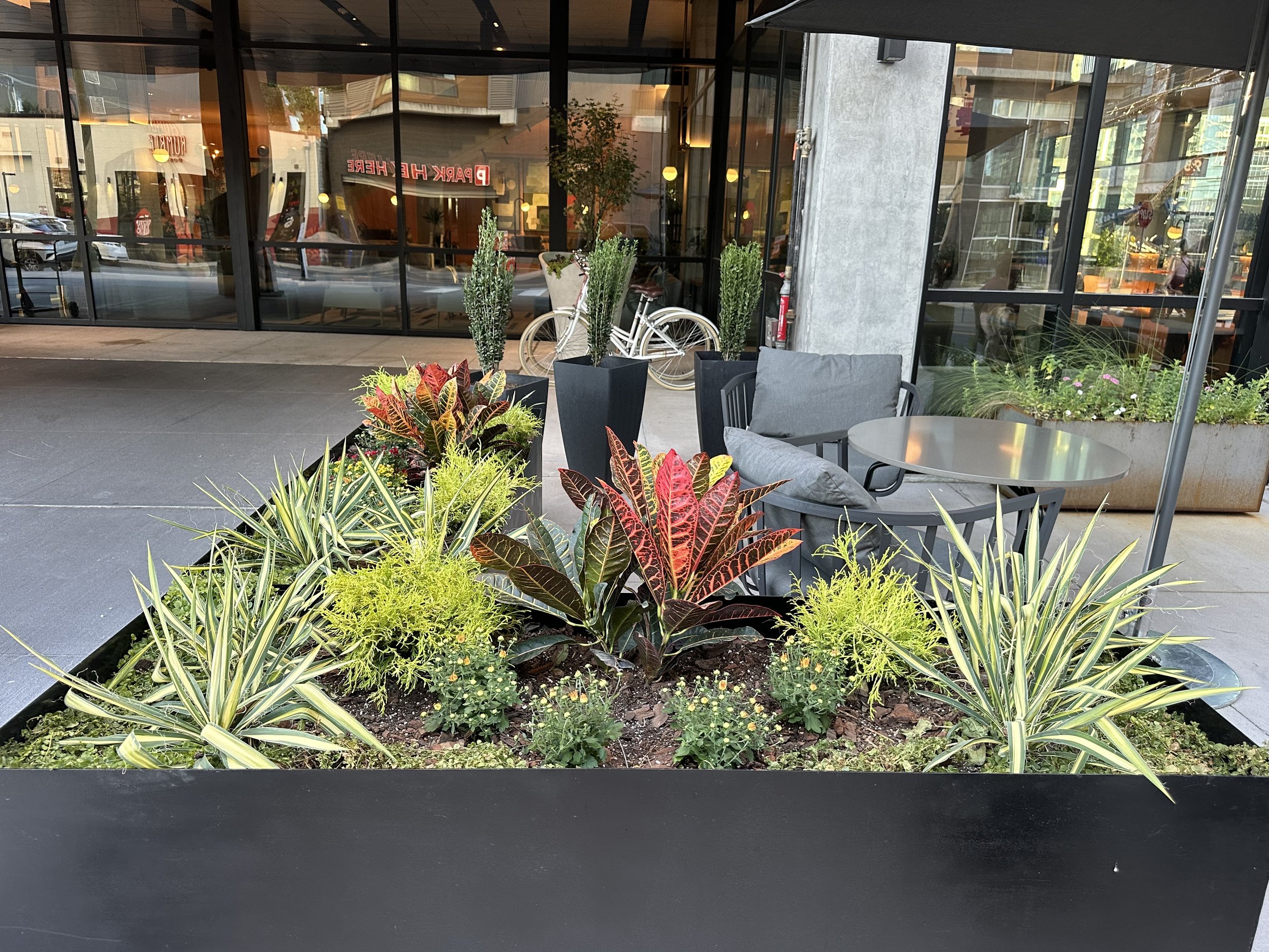 Outdoor seating with a gray sofa, round table, potted plants, and greenery in an urban setting.