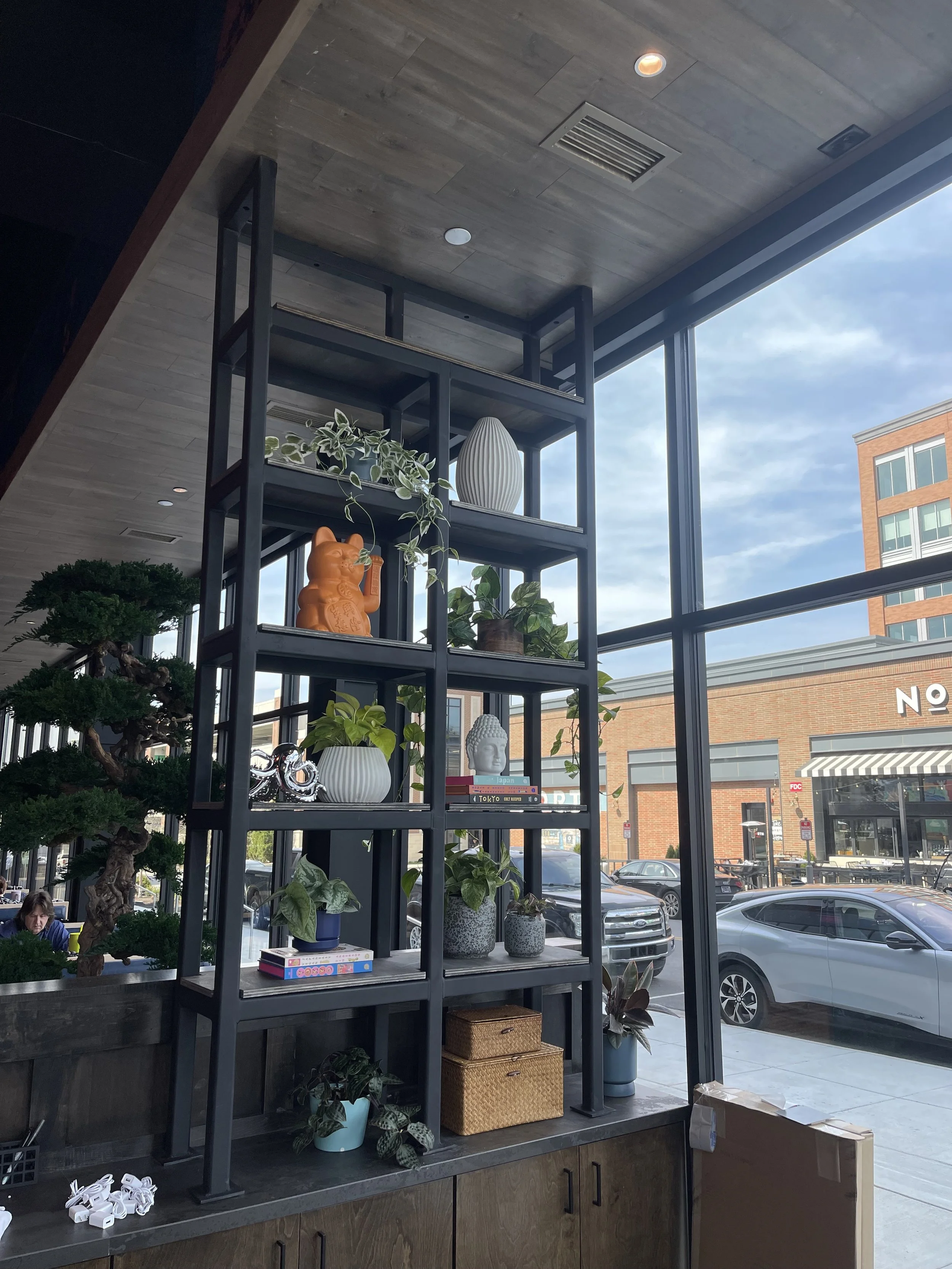 Interior view of a modern storefront with a black metal shelf filled with plants and decorative objects, large window showing parked cars and a brick building outside.