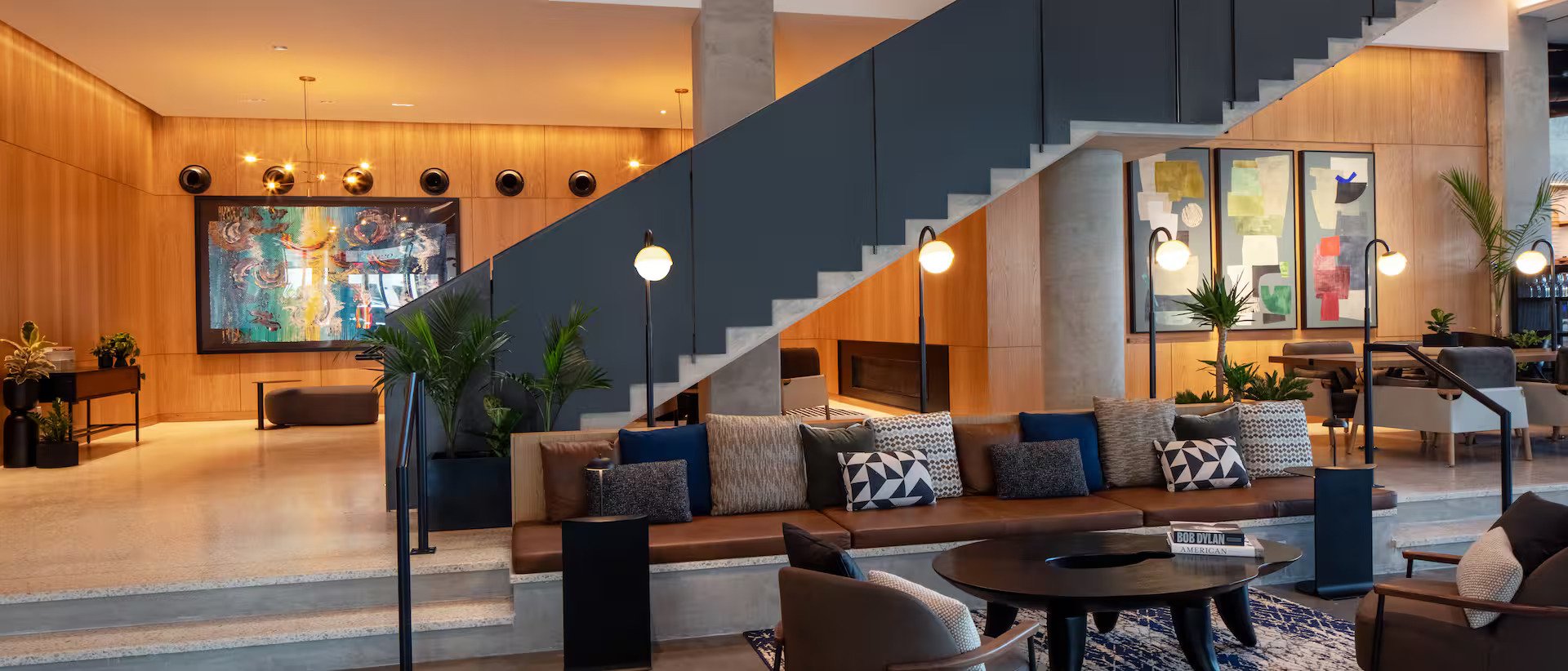 Modern hotel lobby with wooden walls, a staircase with black railing, a large colorful abstract painting, and seating with pillows. There are plants and lamps providing warm lighting.