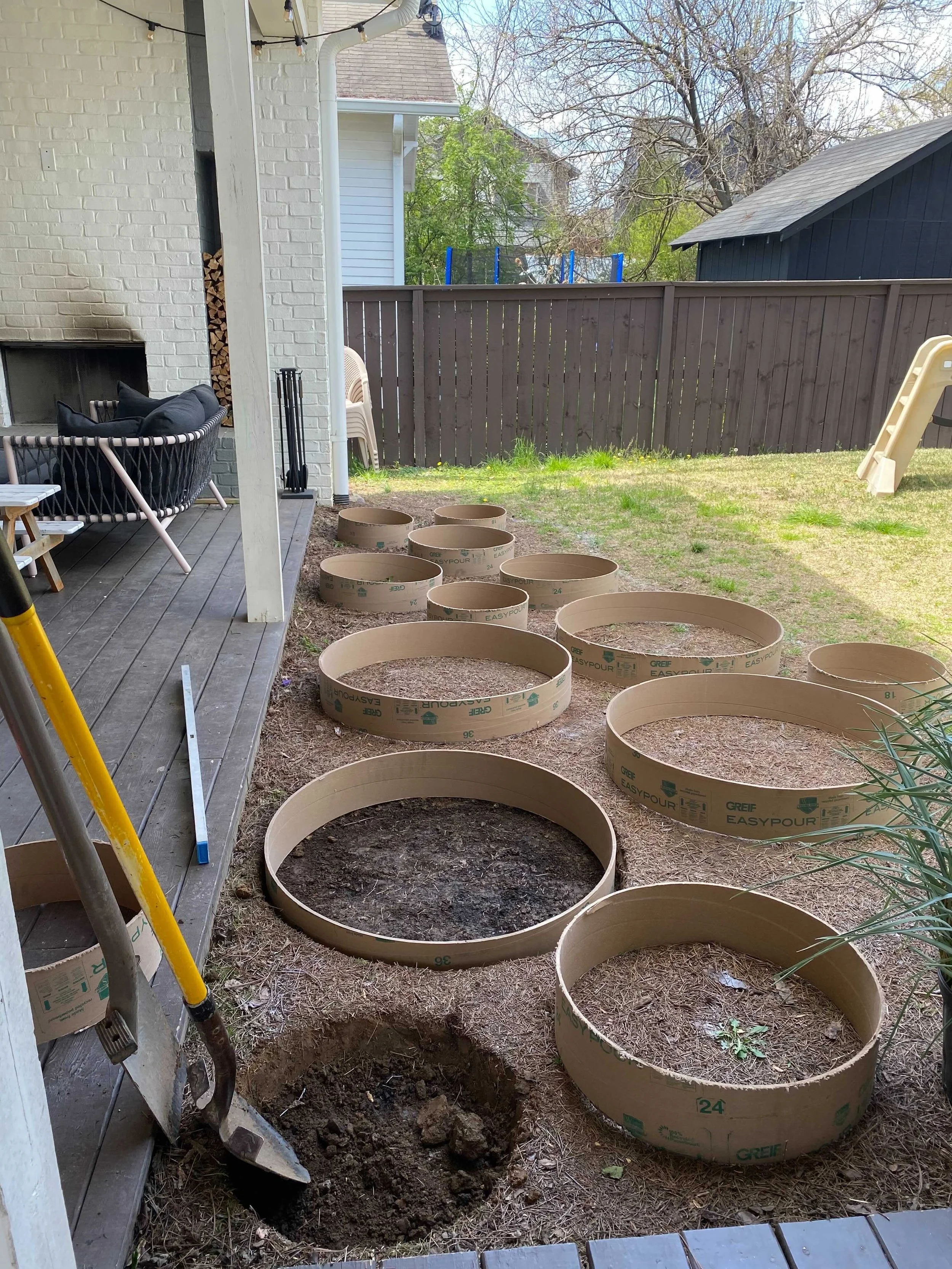 Backyard garden area with cardboard rings laid out on the ground, likely for planting. A shovel is leaning against the house, and there is a patio with furniture and a grill. The yard is fenced, with a small slide in the background.