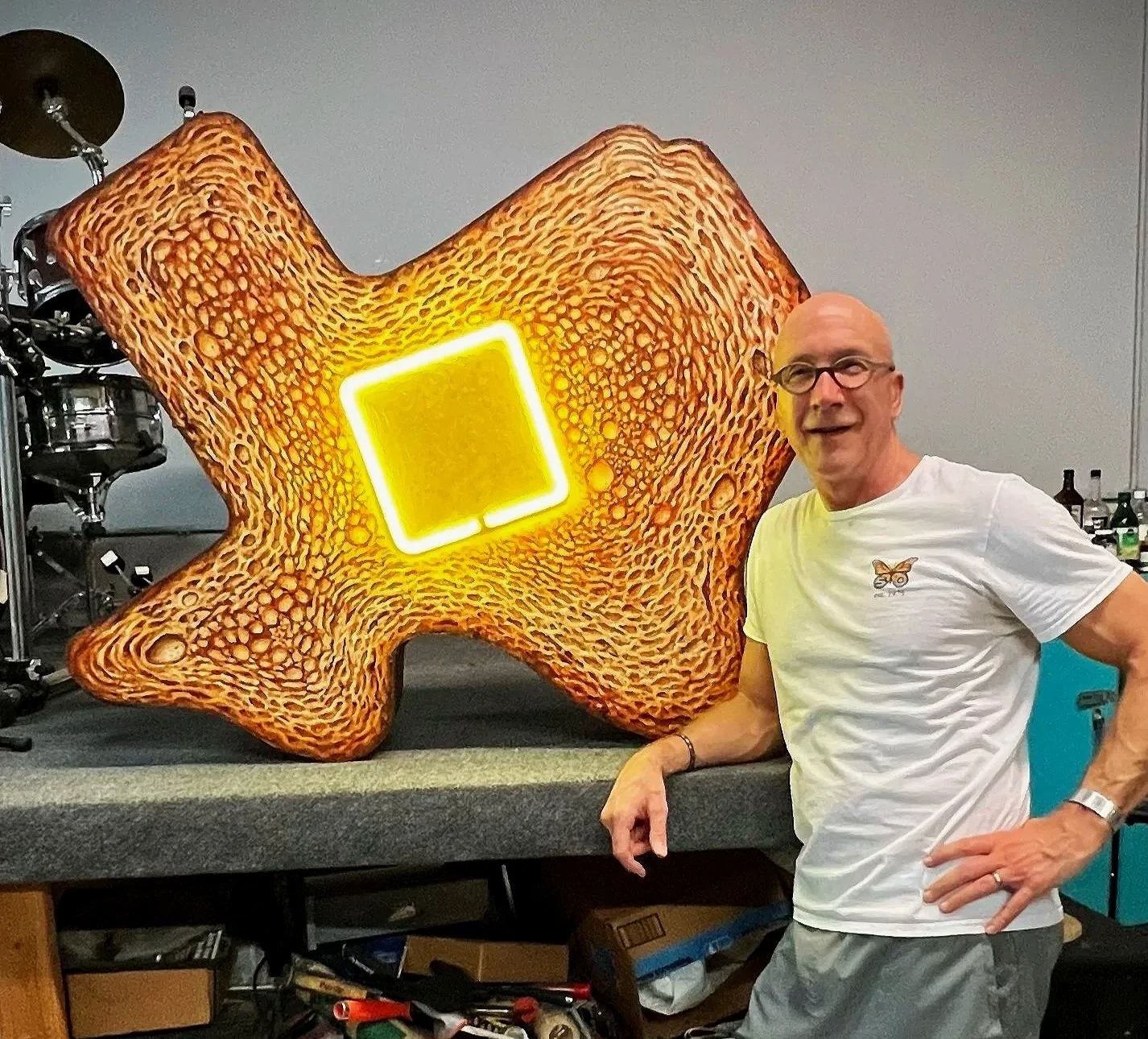 A man with glasses and a white t-shirt standing next to a large, colorful art piece resembling a star-shaped object with a textured surface and a neon yellow square in the center, in a room with musical equipment and bottles in the background.