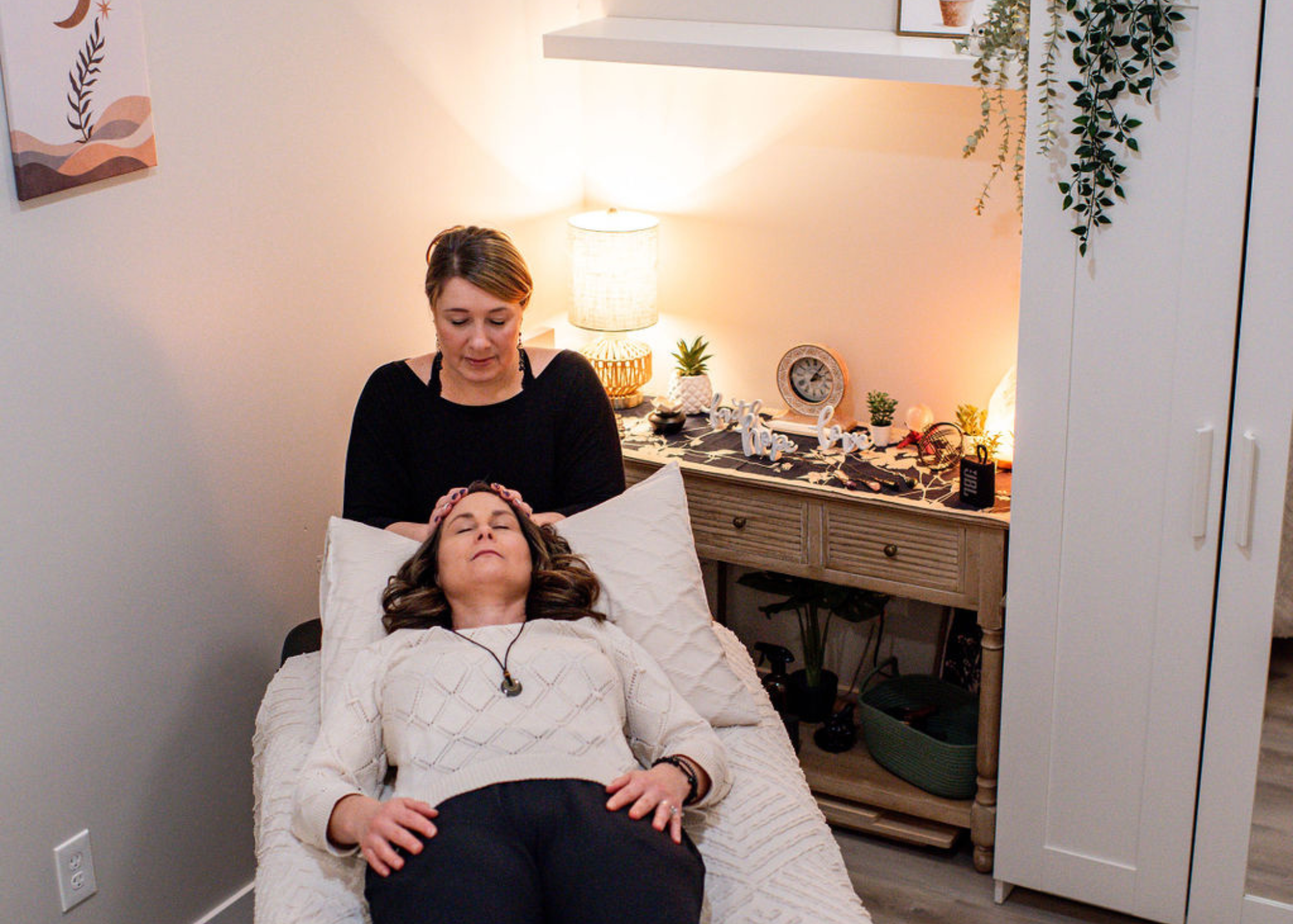 A woman receiving a reiki healing session from Jenie Coaches in a cozy, decorated room with warm lighting, plants, and decorative items on a wooden side table.