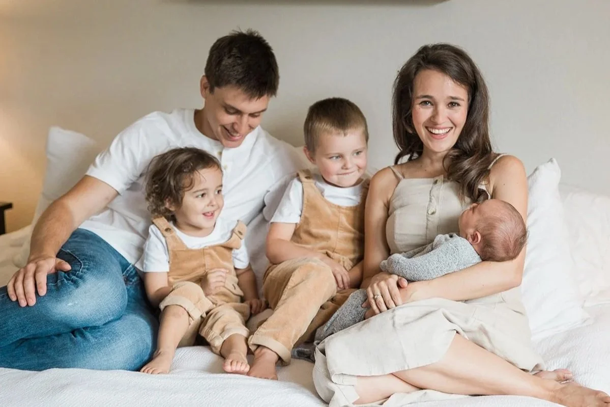 A family sitting on a bed, with a woman holding a newborn baby and smiling, surrounded by two young children and a man.