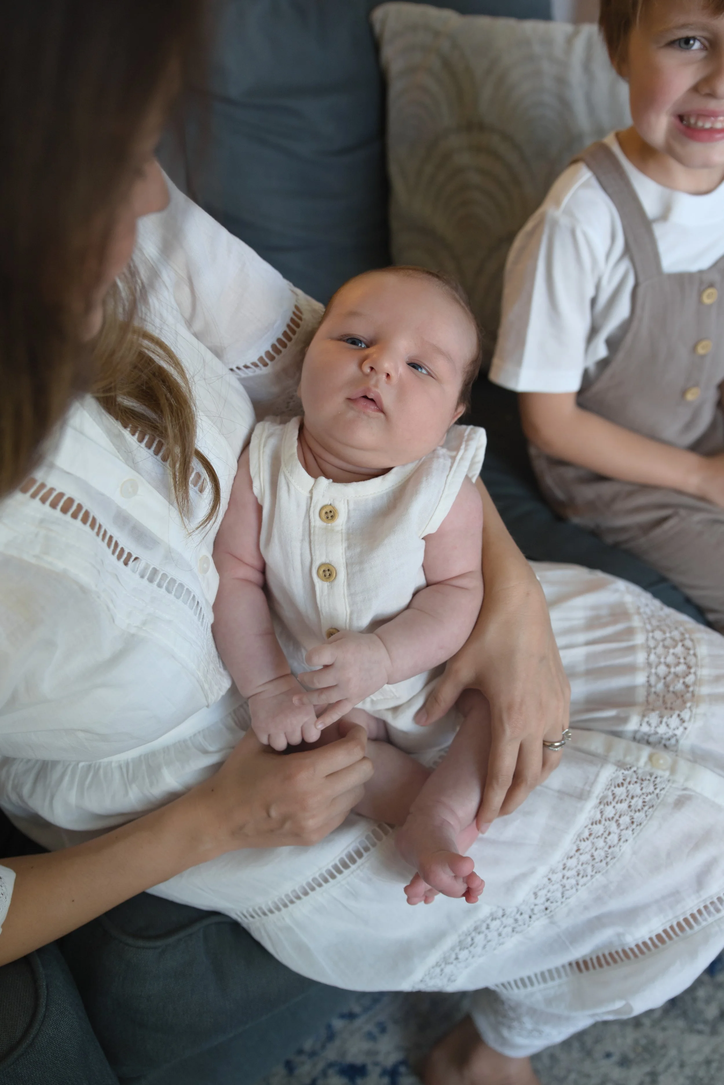 A baby being held by a woman with long, wavy brown hair, sitting on a woman's lap. The baby has light skin, blue eyes, and is wearing a cream-colored sleeveless outfit with buttons. A young girl with light skin, blond hair, and wearing a beige dress with straps and gold buttons is sitting beside them, smiling. They are sitting on a dark-colored sofa with patterned pillows, in a cozy, well-lit room.