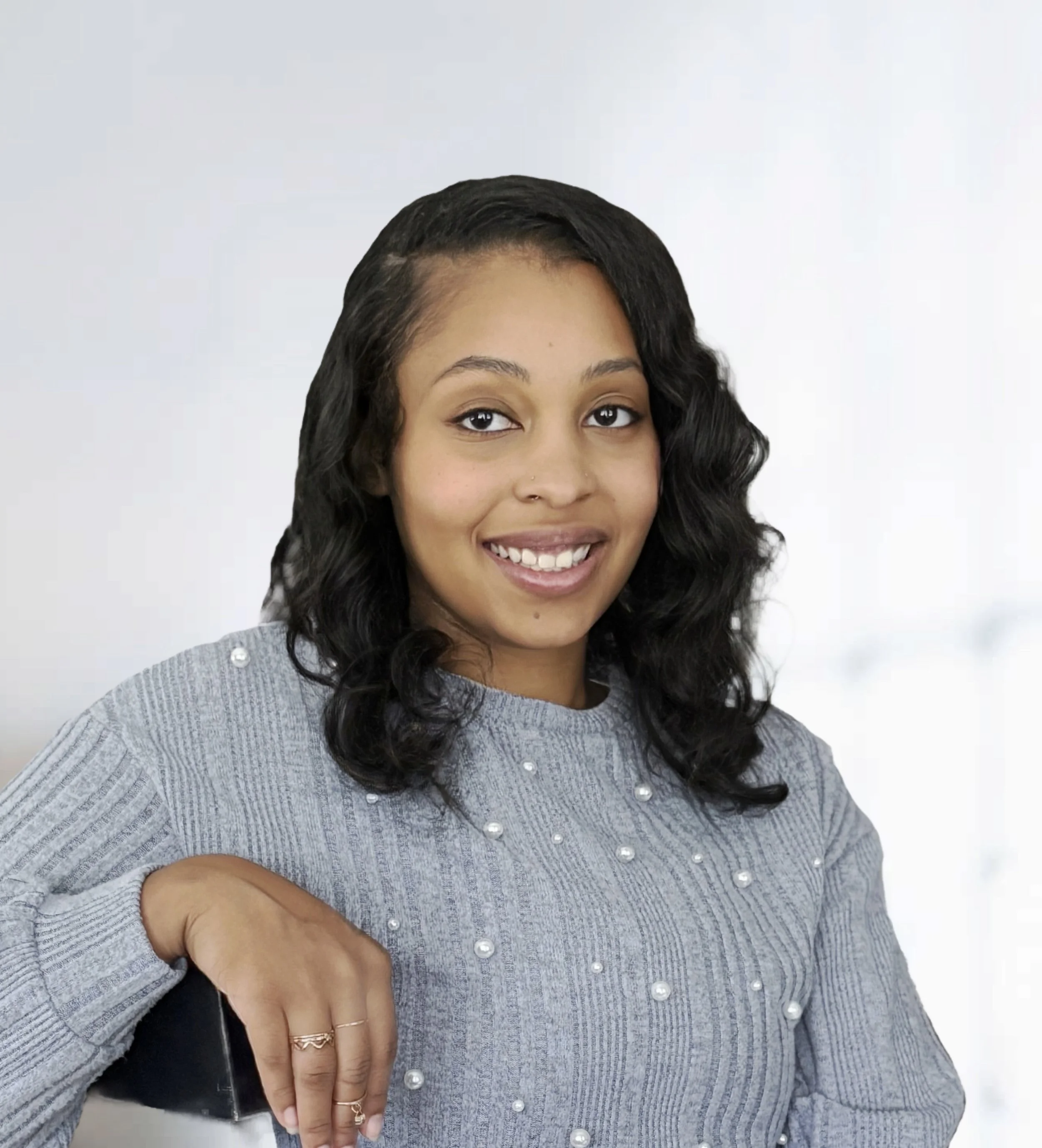 A woman with wavy black hair smiling, wearing a gray sweater with pearl embellishments, leaning on a black chair.
