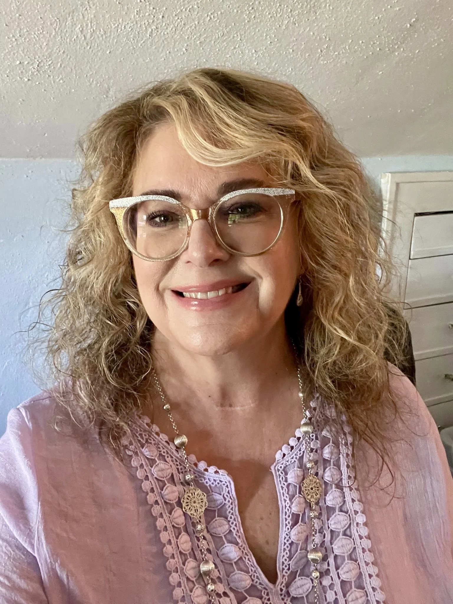A smiling woman with curly blonde hair and glasses, wearing a pink lace top and a beaded necklace, standing in front of a light-colored wall and a white dresser.